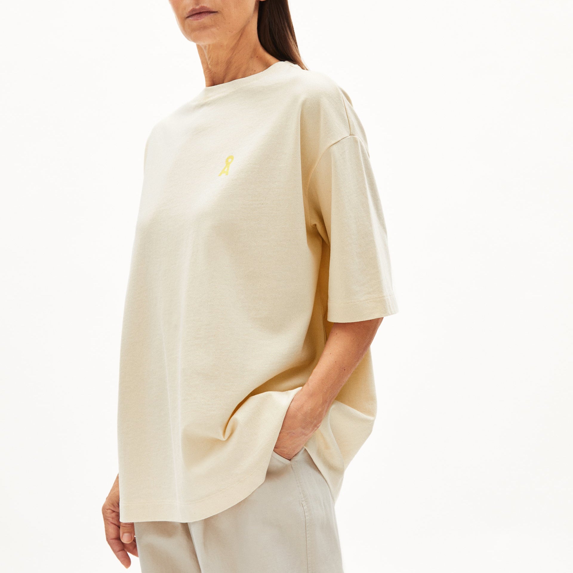 Frau trägt ein beiges T-Shirt mit kleinem gelbem Logo und beigen Hosen, vor einem weißen Hintergrund.