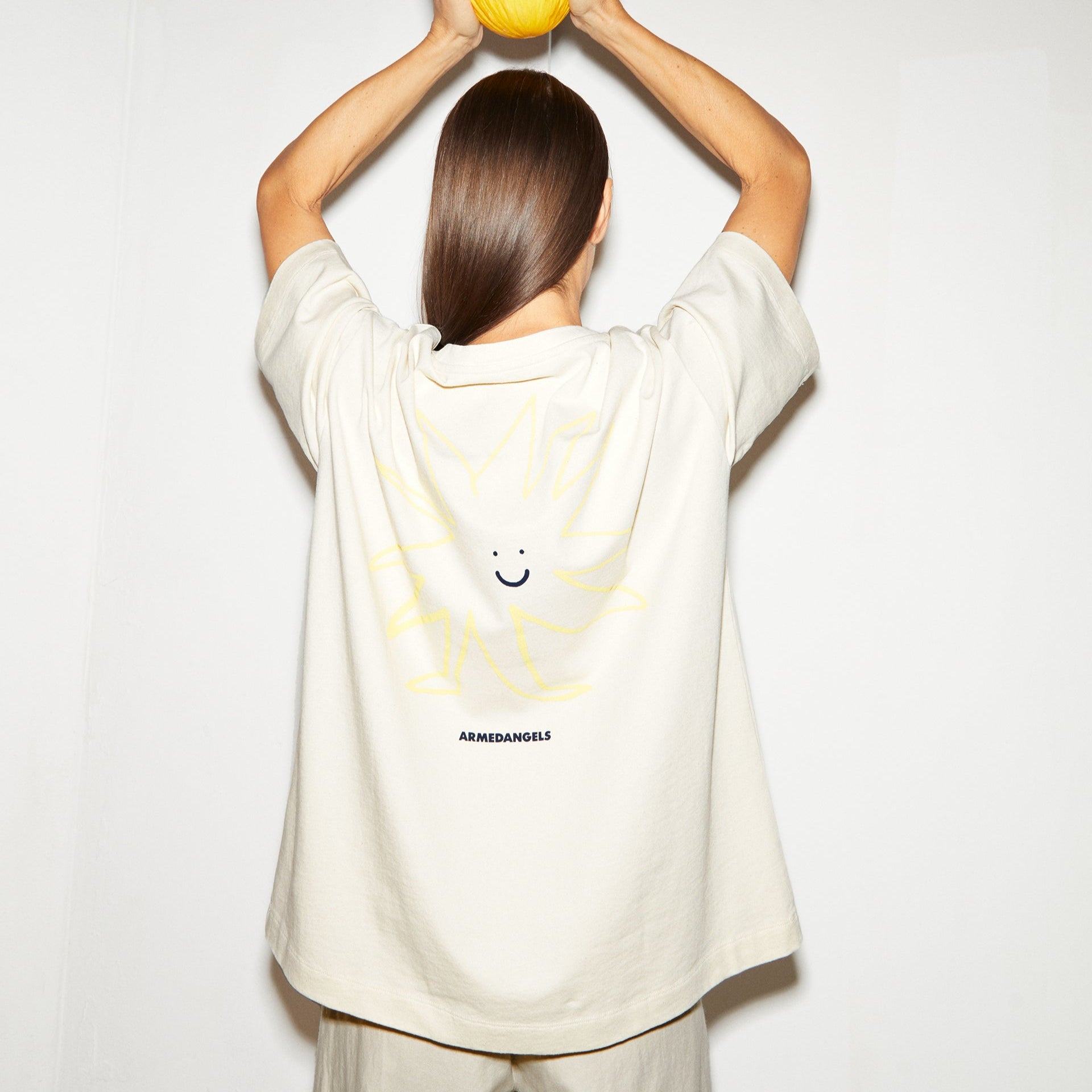 Eine Person hält einen gelben Ball über dem Kopf und trägt ein T-Shirt mit einem lächelnden Sonnengesicht auf dem Rücken.