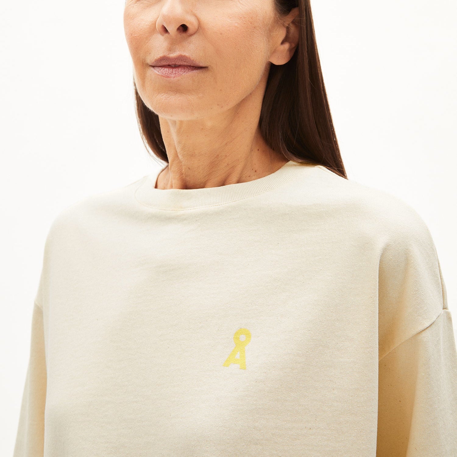 Frau in beigem Oberteil mit gelbem Logo auf der Brust vor neutralem Hintergrund.