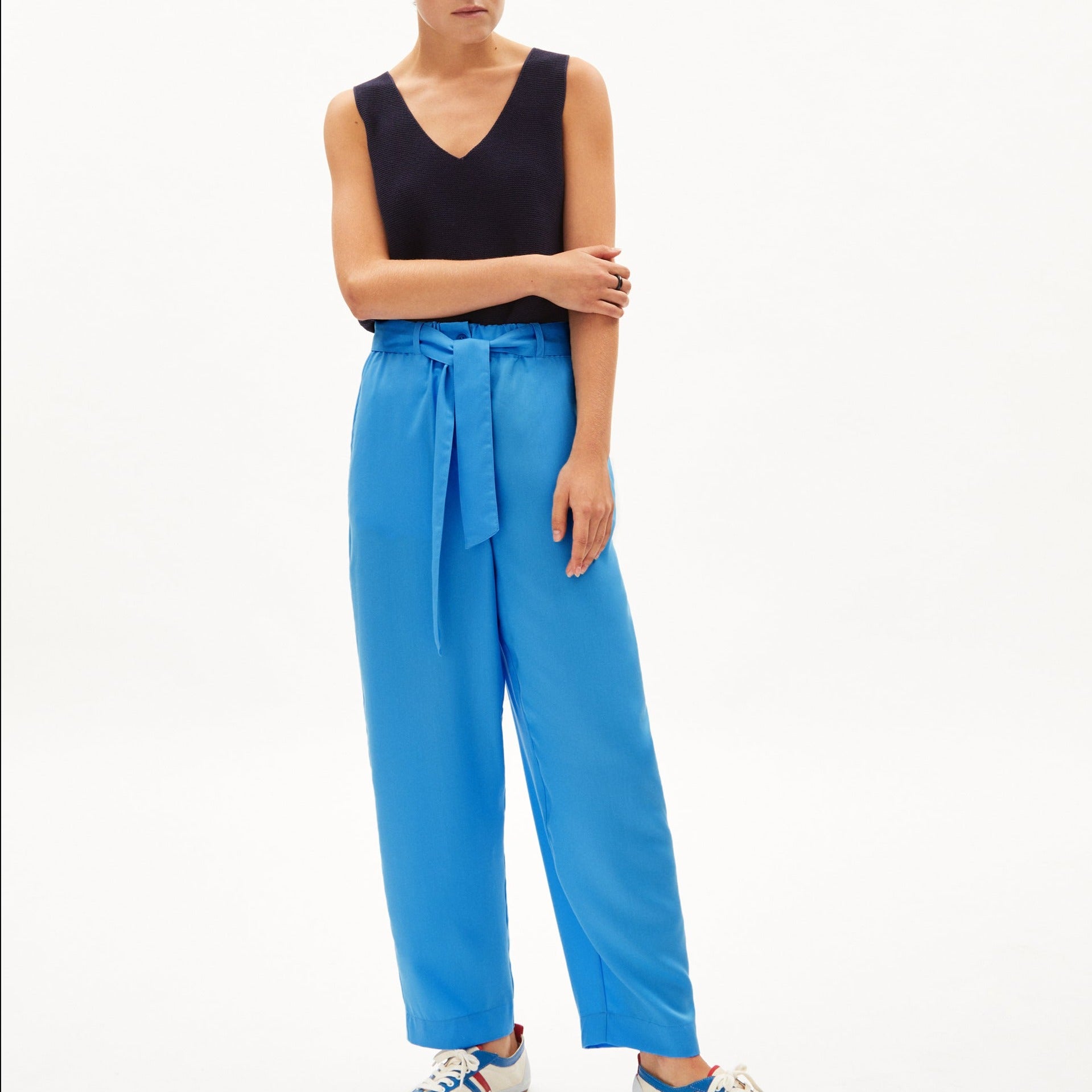 Person in einem ärmellosen schwarzen Oberteil und blauer Hose mit Gürtel, trägt bunte Turnschuhe vor einem weißen Hintergrund.