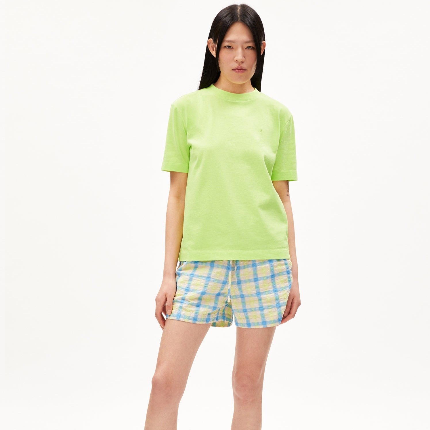 Frau in grünem T-Shirt und karierter Shorts steht vor weißem Hintergrund.