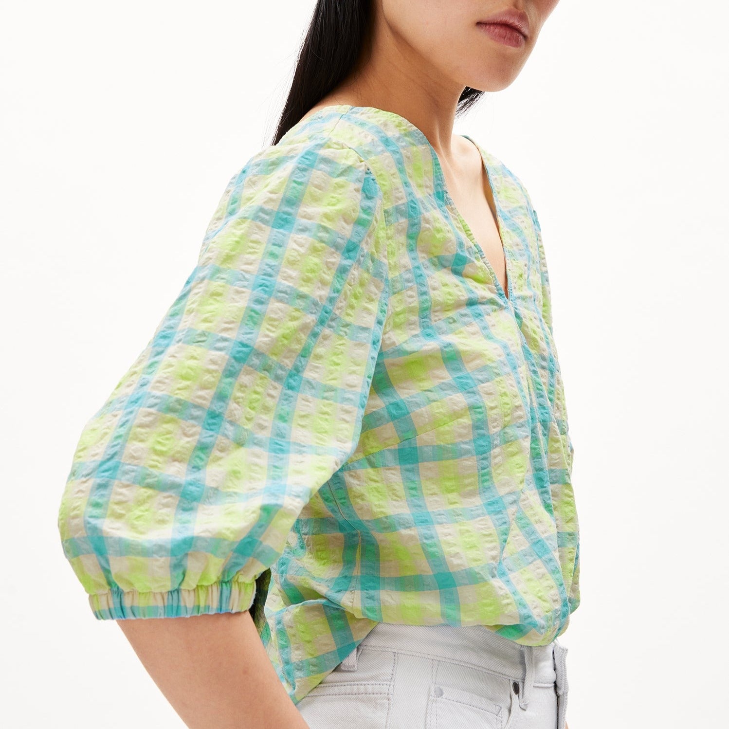 Person in a colorful, checkered blouse with a light blue and green pattern, wearing white pants, facing right.