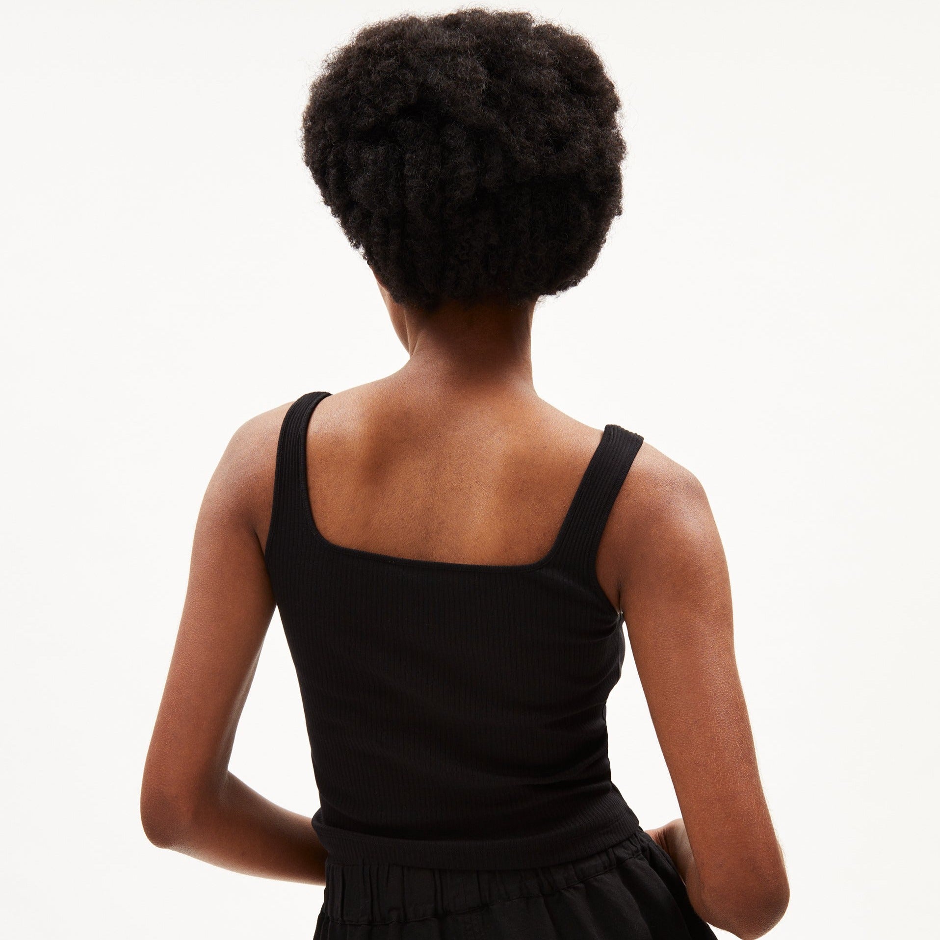 Person von hinten in schwarzem Trägershirt, mit lockigem Haar vor weißem Hintergrund.