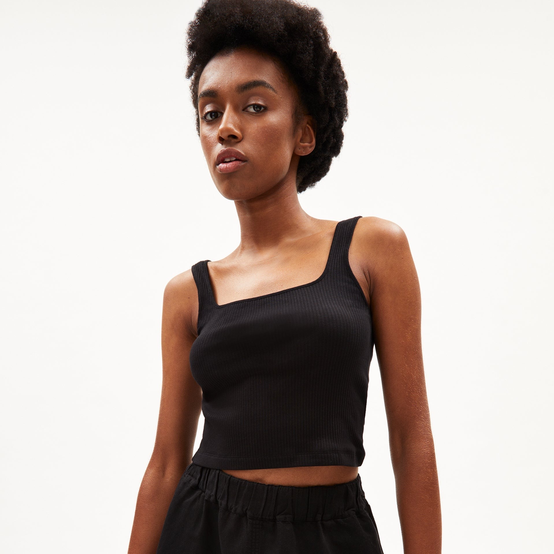 Person mit Afrofrisur trägt ein schwarzes, ärmelloses Top vor weißem Hintergrund.