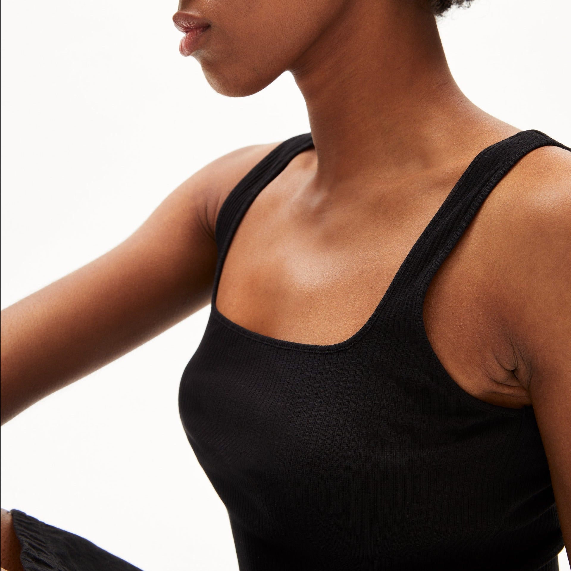 Frau in einem ärmellosen schwarzen Top sitzt, vor weißem Hintergrund, seitlich zur Kamera.