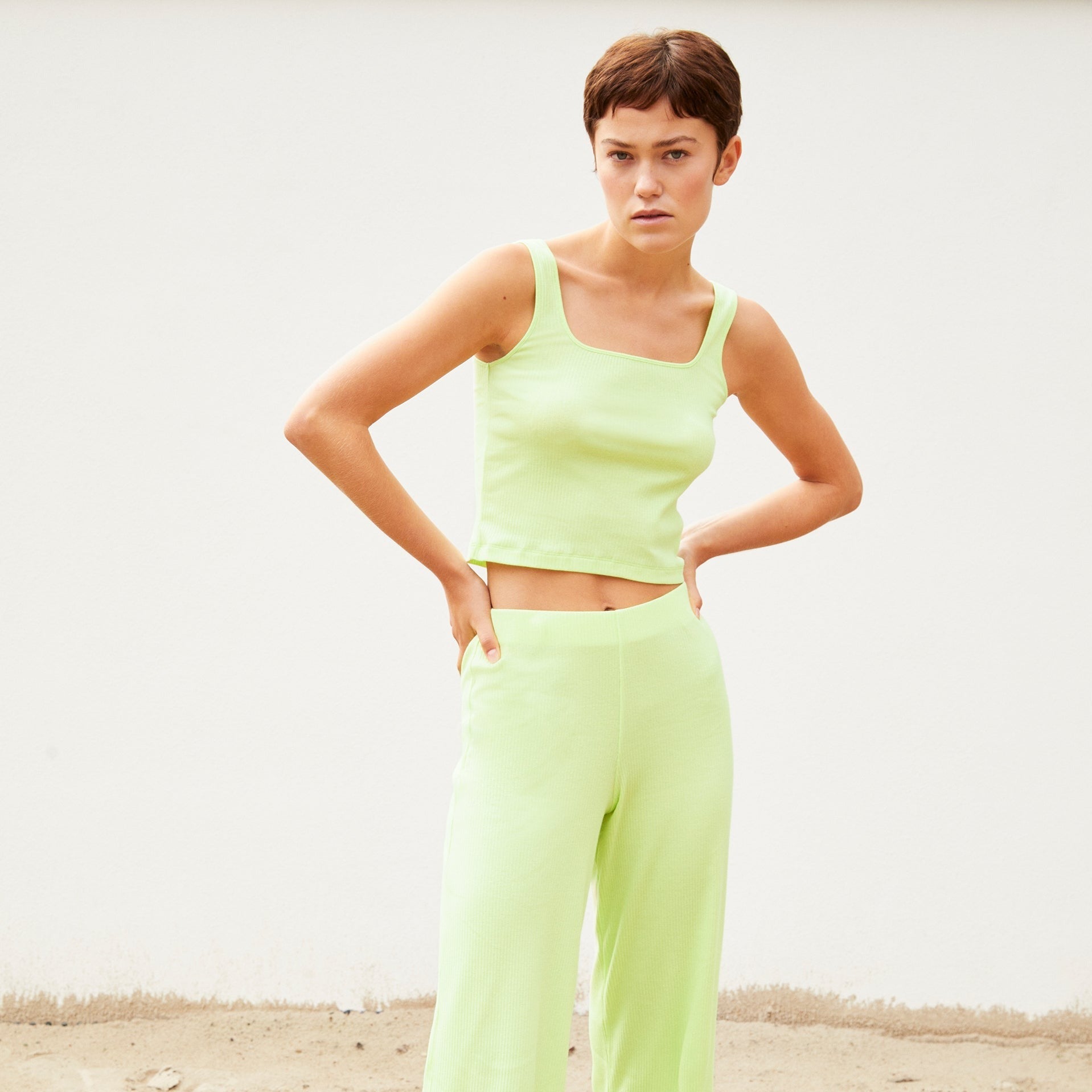 Person in grünem Outfit steht vor einer weißen Wand mit ernster Miene.