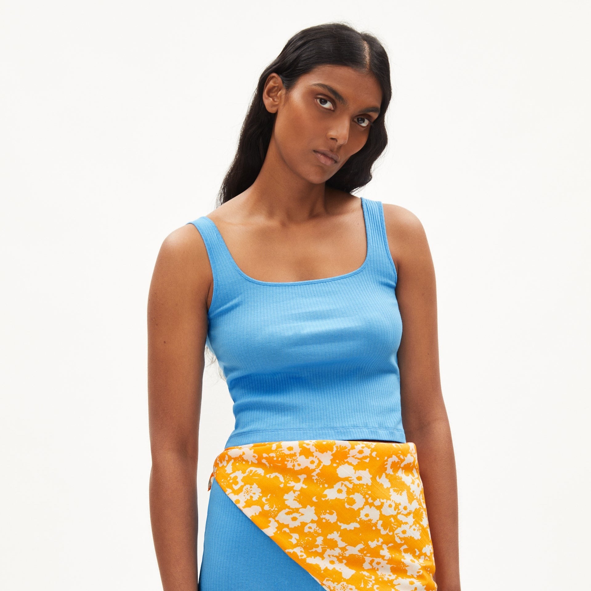 Frau in blauem Top und gelb-gemustertem Rock steht vor neutralem Hintergrund.