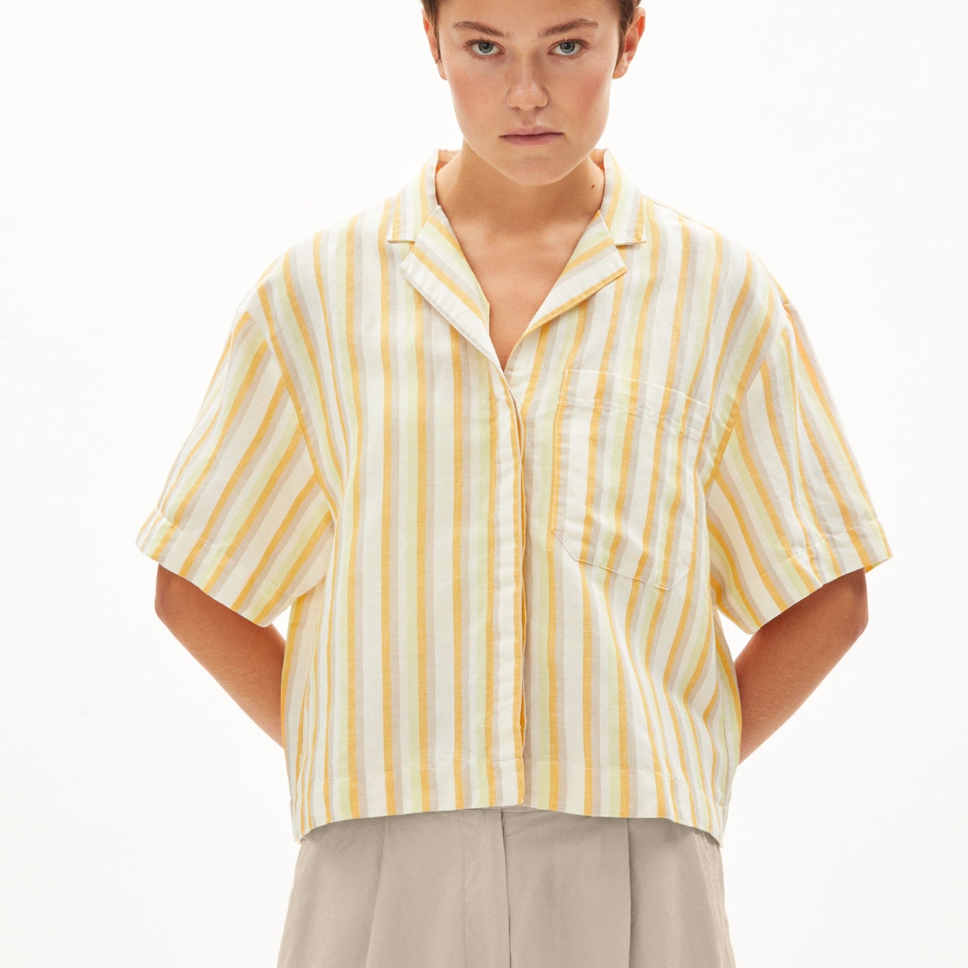 Eine Person trägt ein gelb-weiß gestreiftes Hemd und beige Hose vor einem weißen Hintergrund.