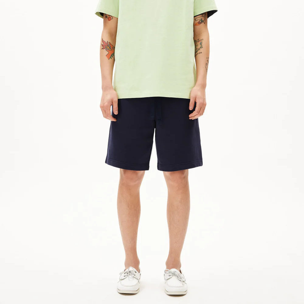 Eine Person mit rasiertem Kopf und sichtbaren Armtattoos steht mit dem Gesicht nach vorne, trägt ein hellgrünes T-Shirt, marineblaue Shorts und weiße Turnschuhe vor einem schlichten weißen Hintergrund.