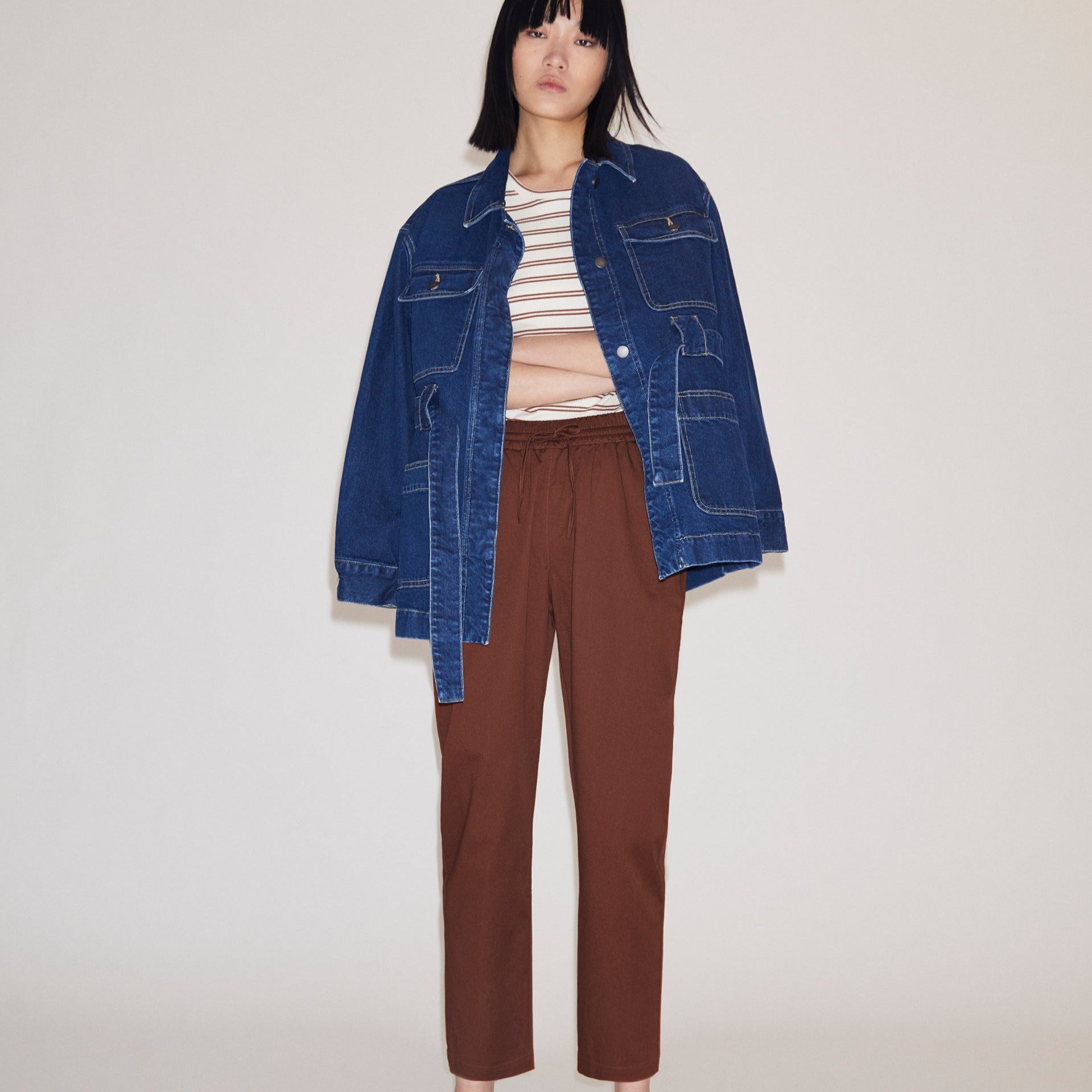 Frau mit blauer Jeansjacke, gestreiftem Oberteil und brauner Hose posiert vor neutralem Hintergrund.
