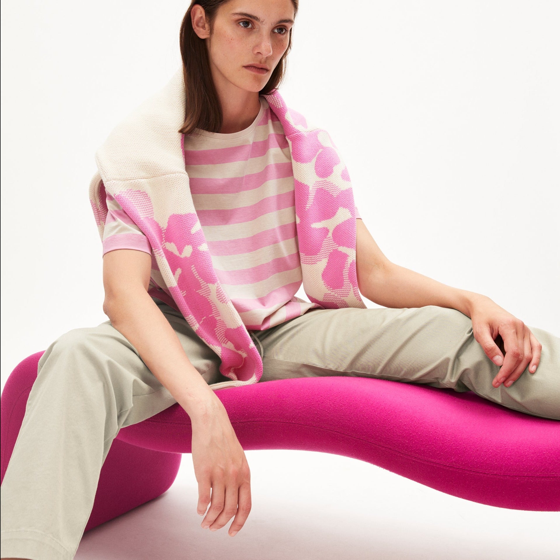 Person auf pinkem, gebogenem Stuhl, trägt gestreiftes Shirt und Jacke über den Schultern.