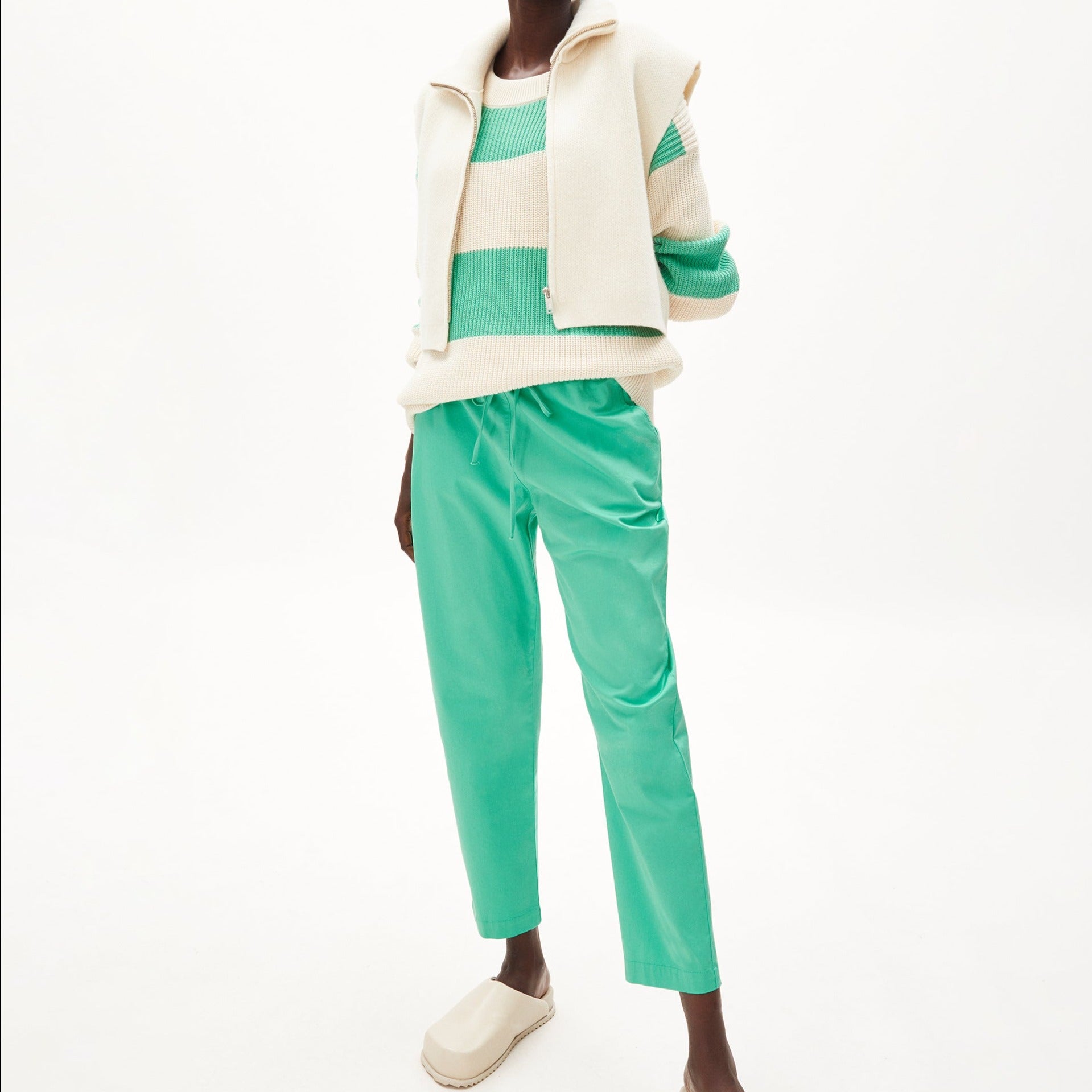 Person in grün-weiß gestreiftem Pullover, grüner Hose und cremefarbener Weste vor weißem Hintergrund.