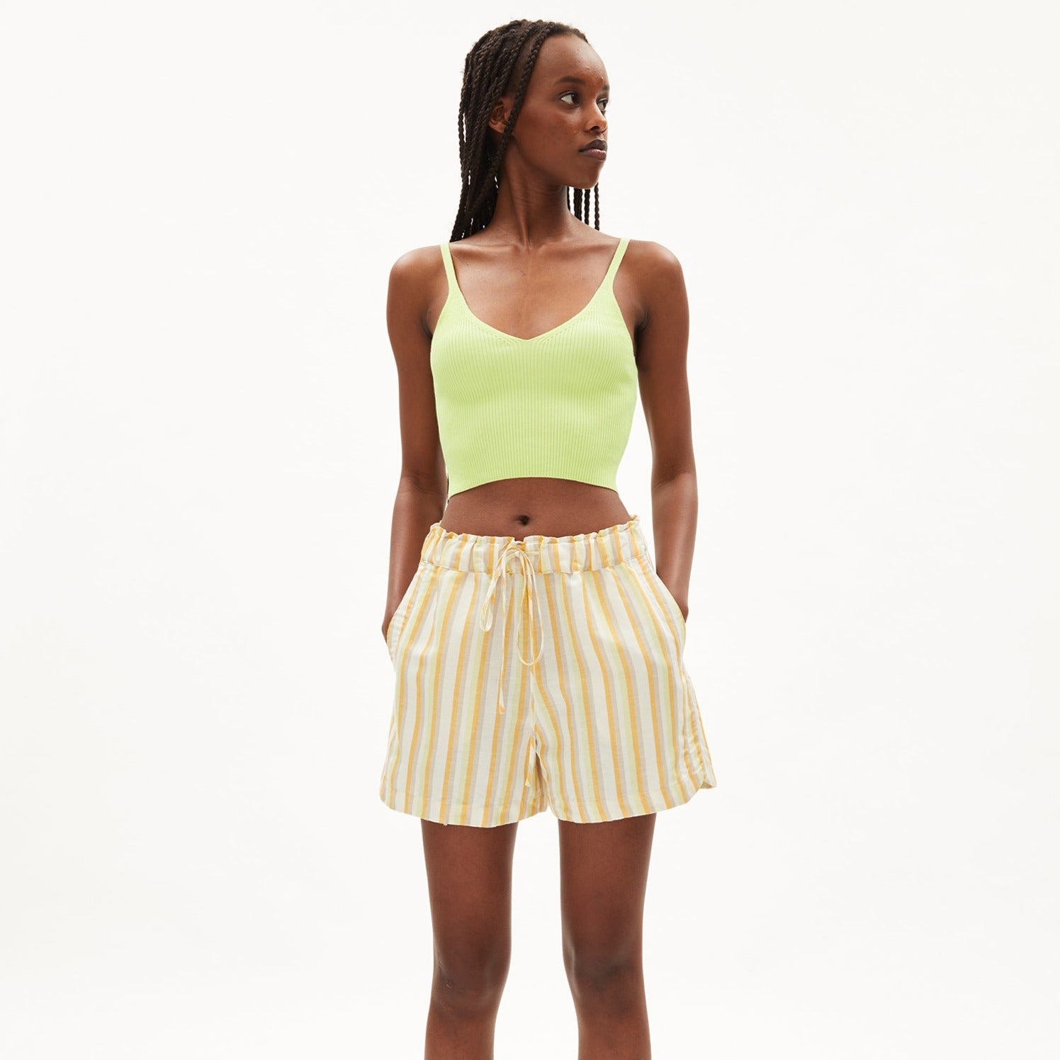 Eine Person trägt ein hellgrünes Crop-Top und gestreifte gelb-weiße Shorts, steht vor einem weißen Hintergrund.