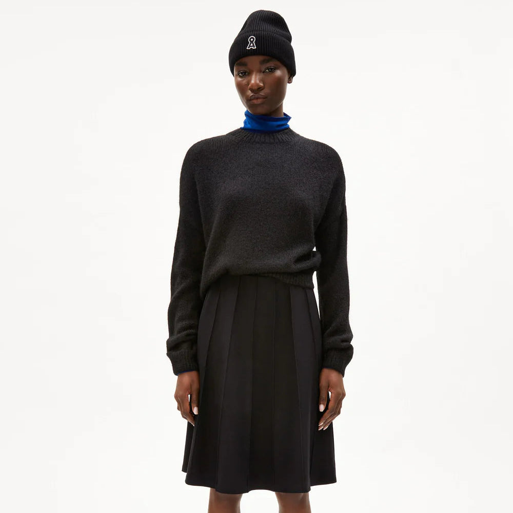 Eine Person steht vor einem schlichten weißen Hintergrund und trägt eine schwarze Mütze, einen dunklen Pullover, einen blauen Rollkragenpullover, einen schwarzen Faltenrock und schwarze Schuhe mit Schnallen.