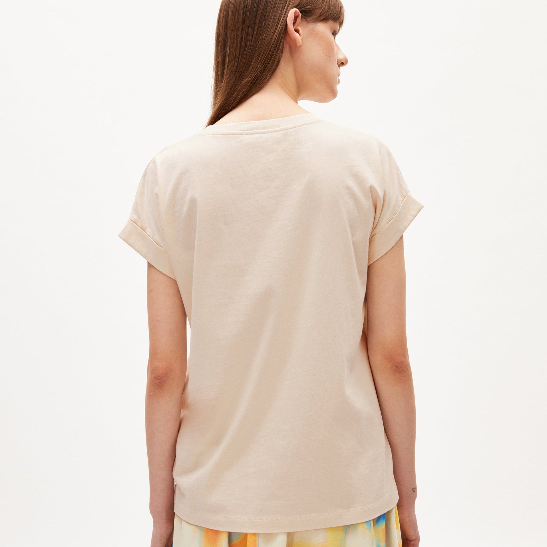 Eine Person mit langen Haaren trägt ein beigefarbenes T-Shirt und steht mit dem Rücken zur Kamera.