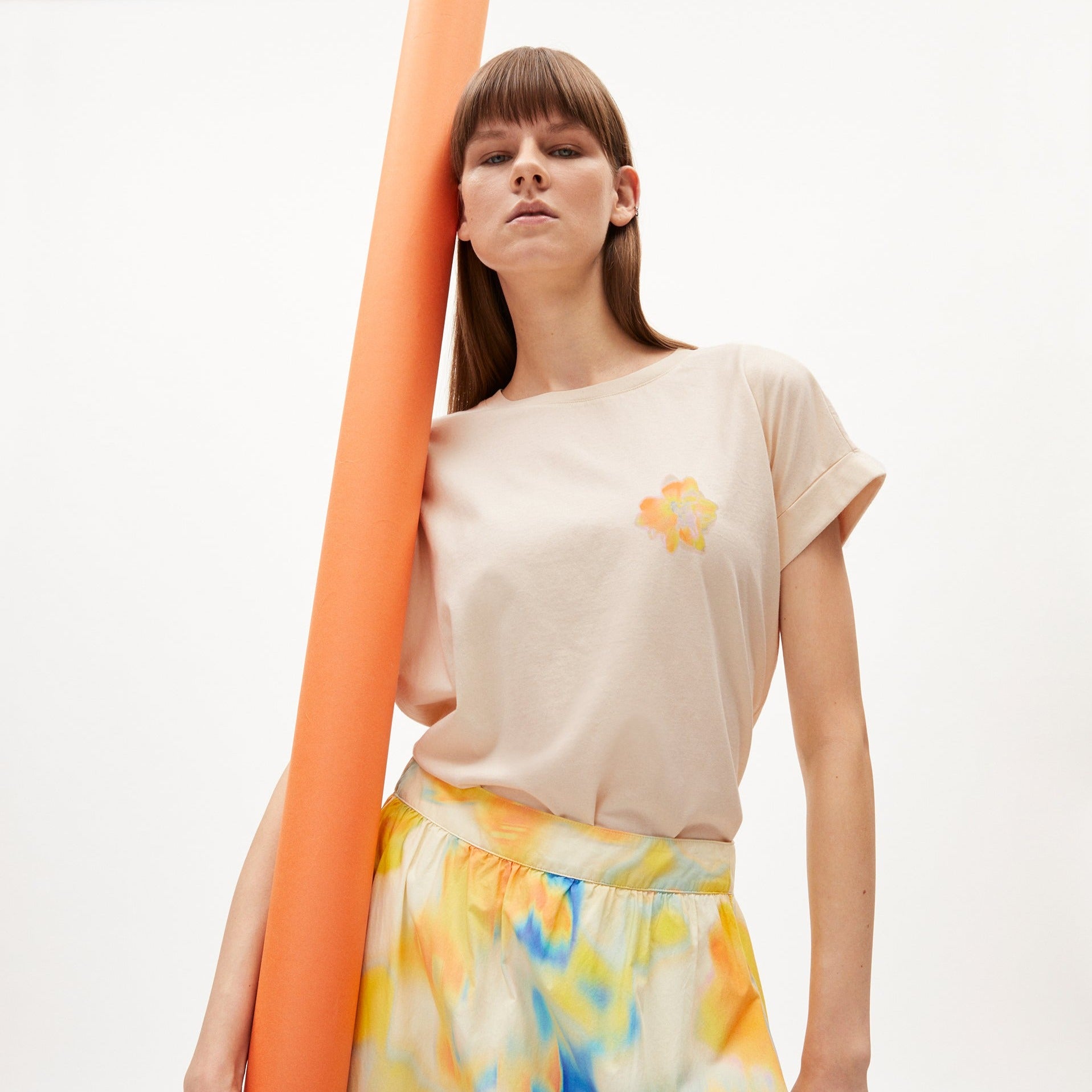 Frau in beigem T-Shirt mit Blumenmuster hält eine große orangefarbene Rolle, trägt einen bunten Rock.
