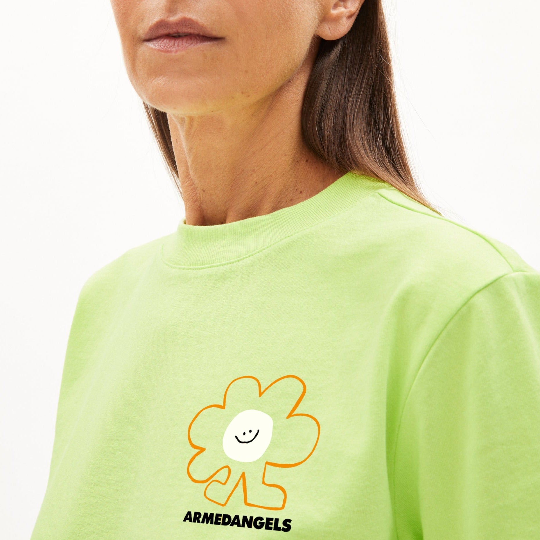 Frau trägt ein hellgrünes T-Shirt mit einem fröhlichen, cartoonhaften Blumenaufdruck und dem Wort "ARMEDANGELS".