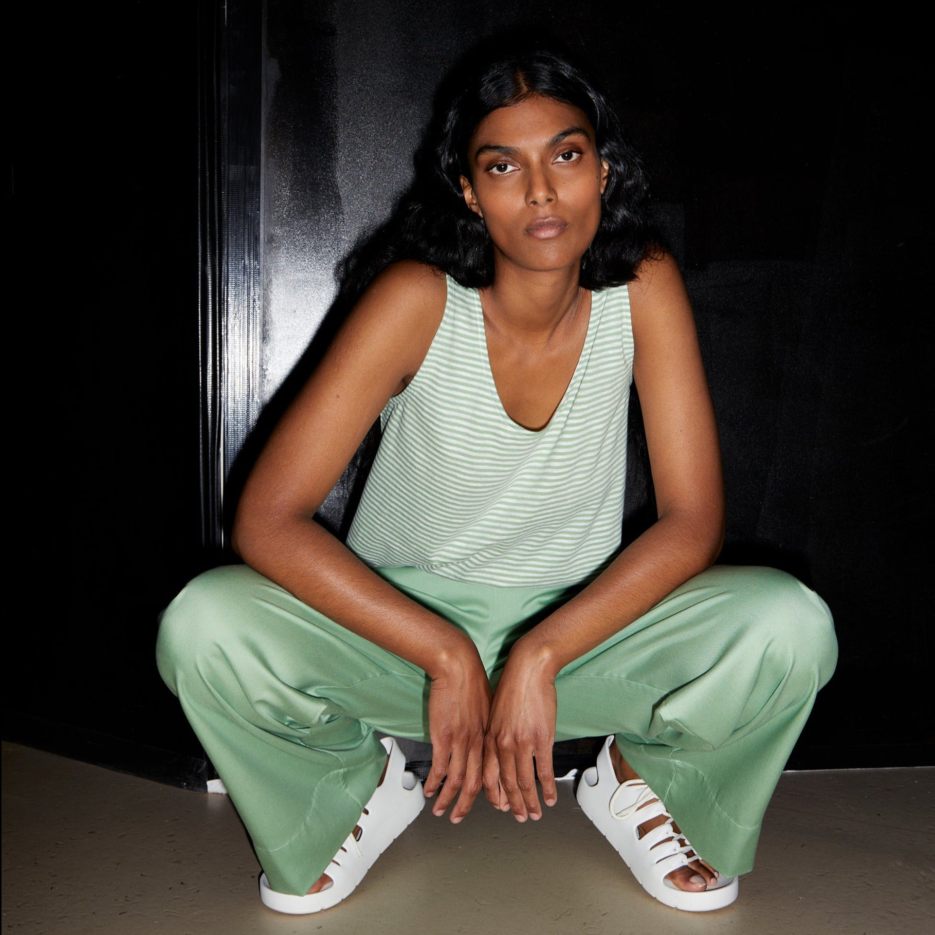 Person in grüner Kleidung hockt vor einem schwarzen Hintergrund und trägt weiße Sandalen.