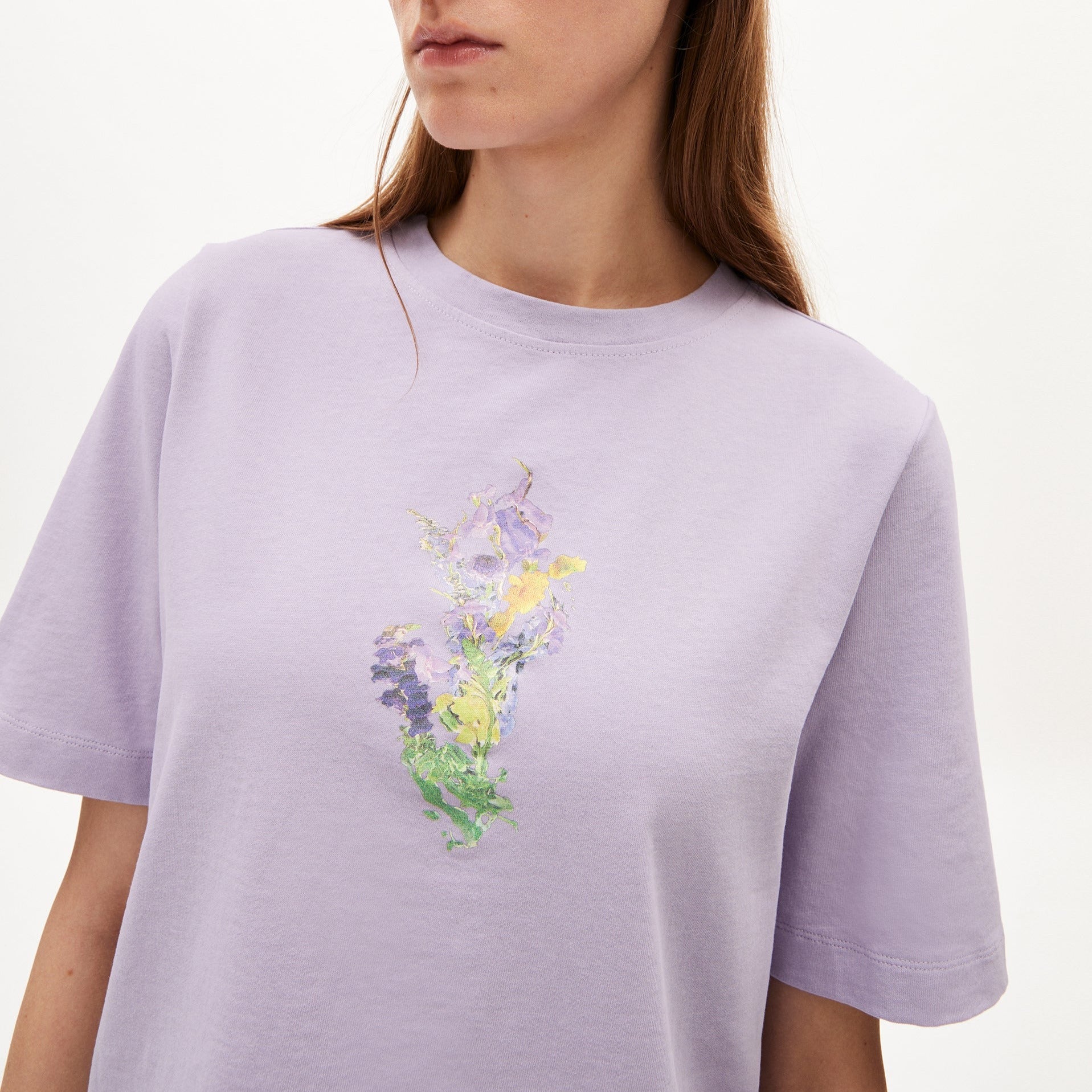 Frau in einem lila T-Shirt mit floralem Design in der Mitte vor neutralem Hintergrund.