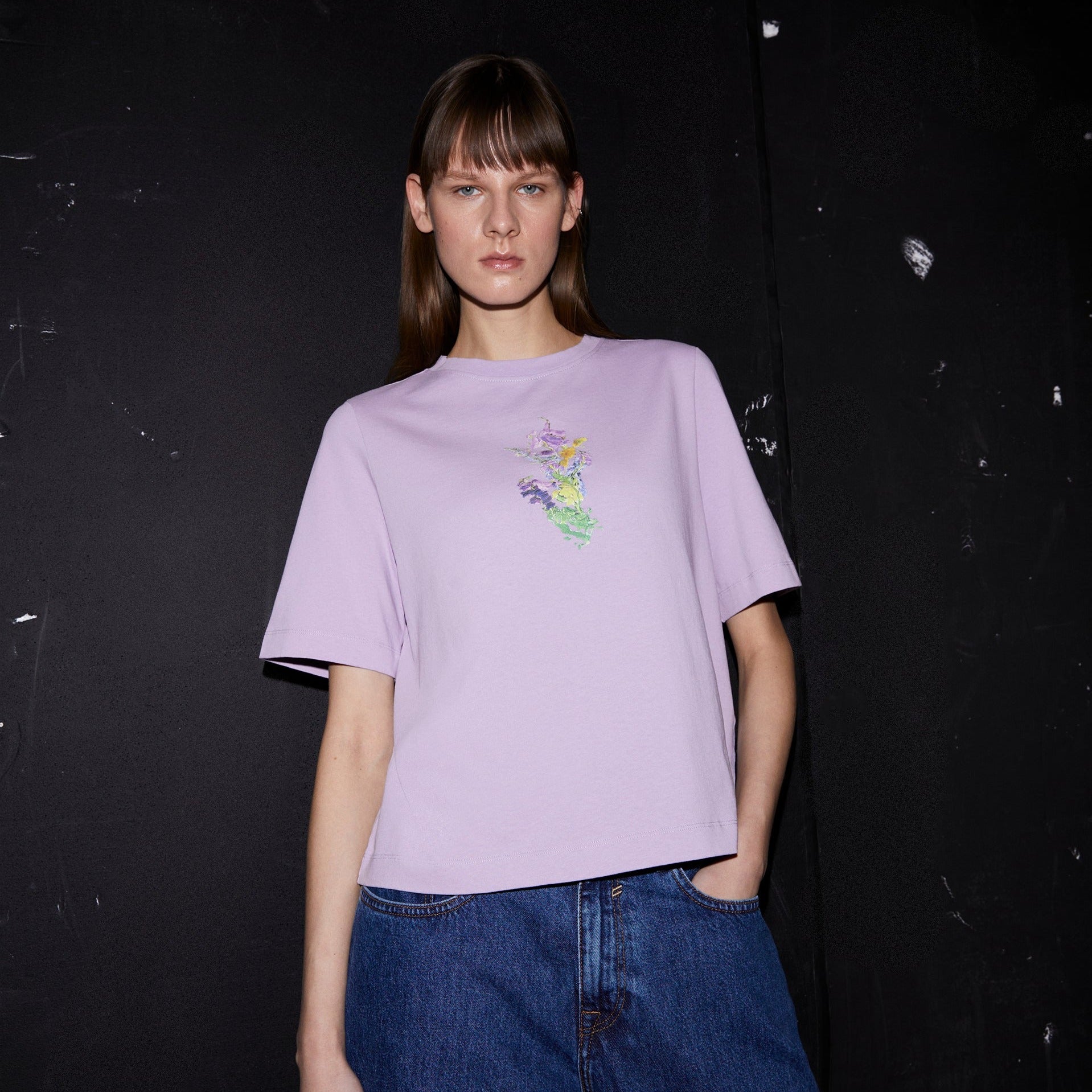 Person in einem lila T-Shirt mit buntem Muster, Jeans, vor schwarzem Hintergrund.