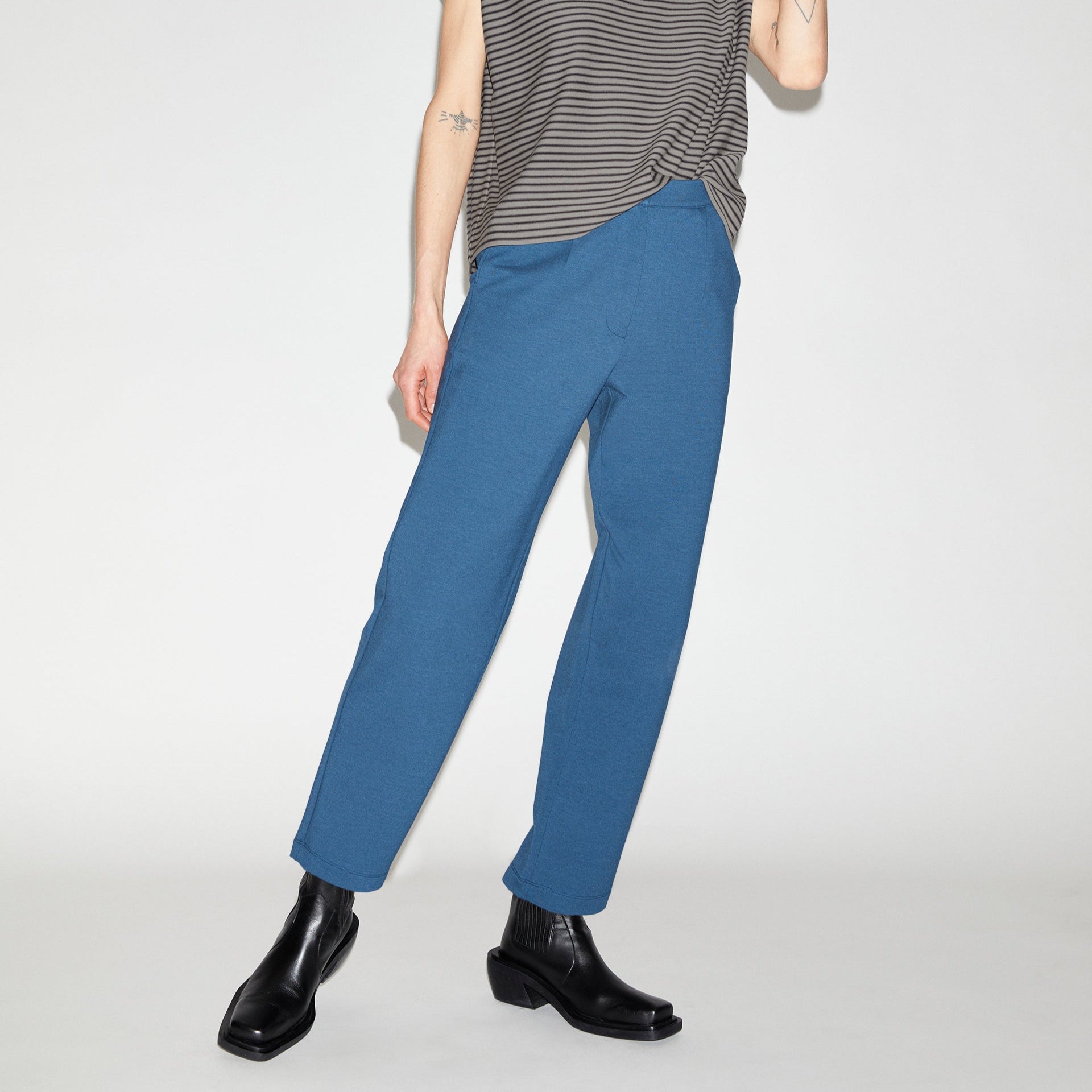 Person in gestreiftem Oberteil und blauer Hose mit schwarzen Stiefeln, steht vor weißem Hintergrund.