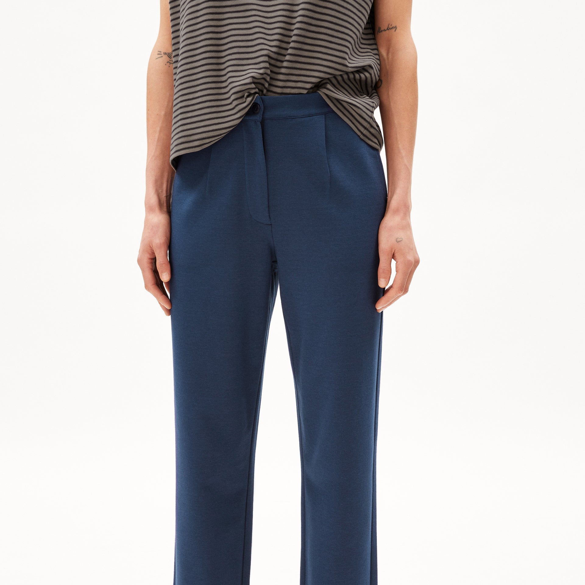 Person in gestreiftem Top und blauer Hose, Fokus auf Körper vom Hals abwärts, neutraler Hintergrund.