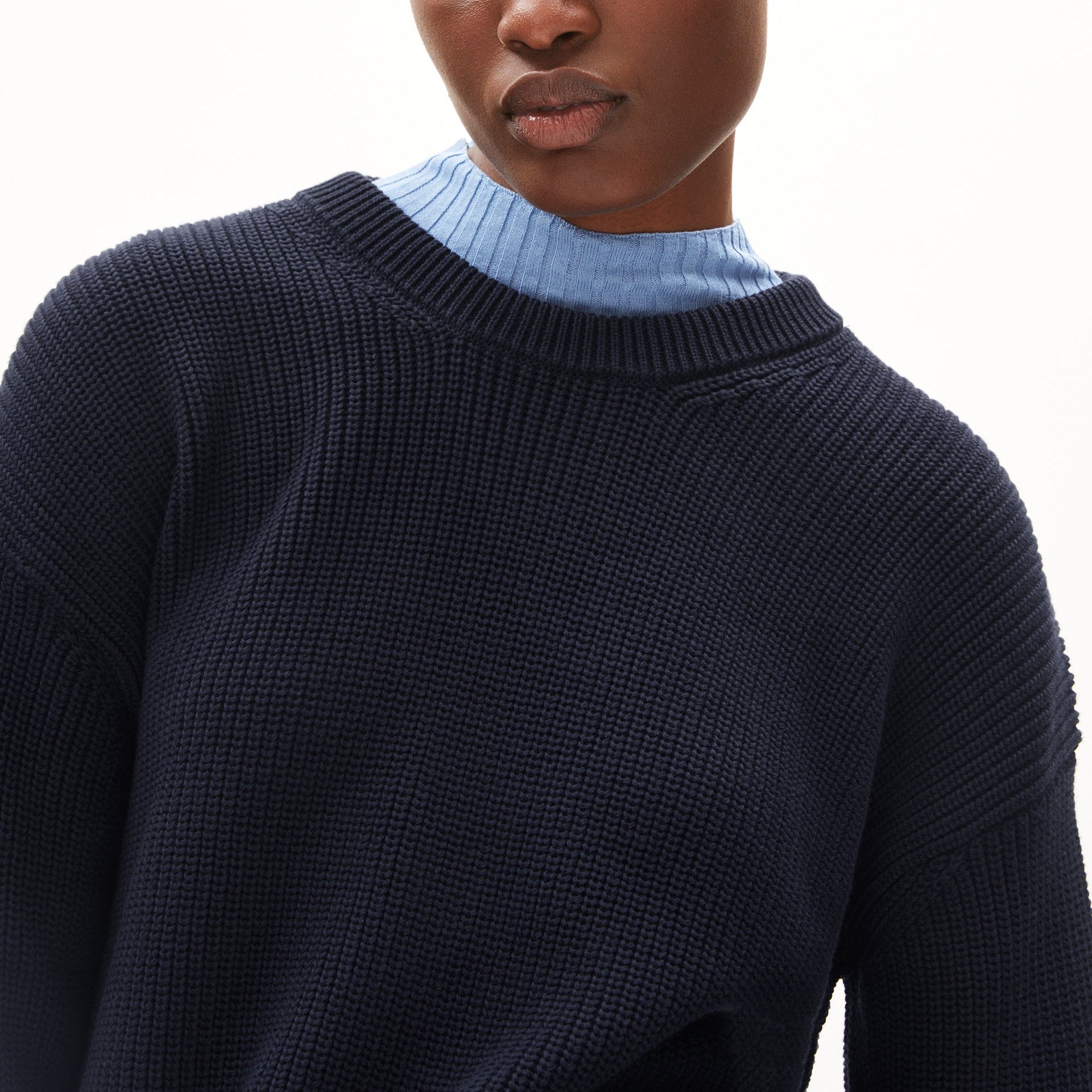 Person in blauem Pullover und hellblauem Oberteil darunter, zeigt Oberkörper und Teil des Gesichts vor weißem Hintergrund.