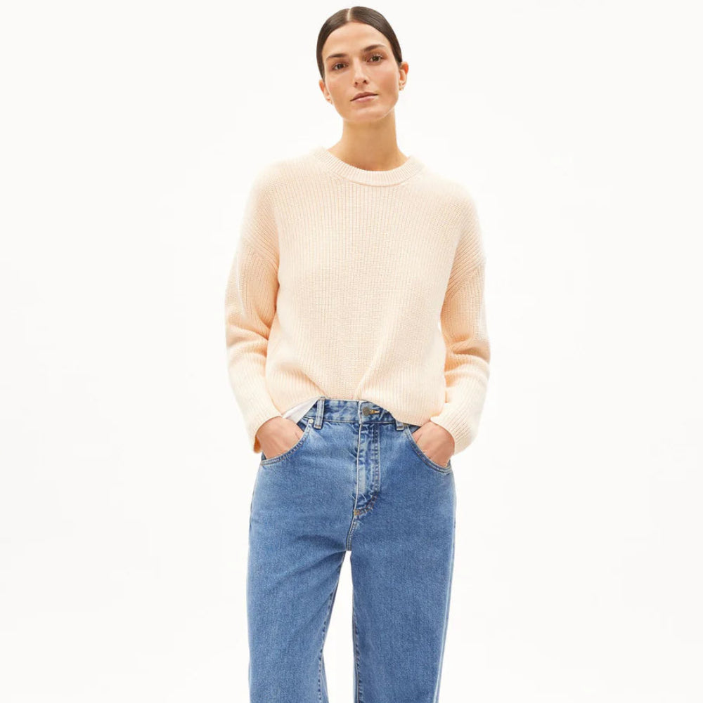 Eine Person mit dunklem, zurückgegeltem Haar, die einen hellbeigen Pullover und blaue Jeans trägt, steht mit den Händen in den Taschen vor einem schlichten weißen Hintergrund.