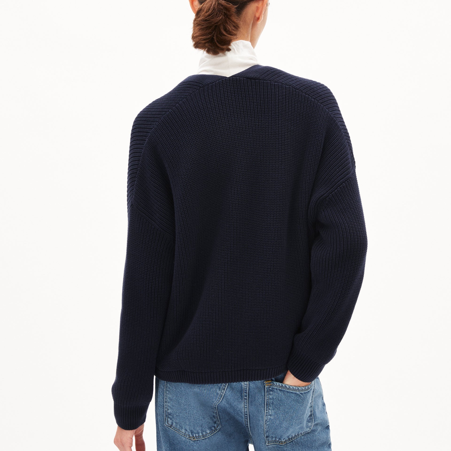 Person mit dunkelblauem Pullover und Jeans von hinten, eine Hand in die Hosentasche gesteckt.