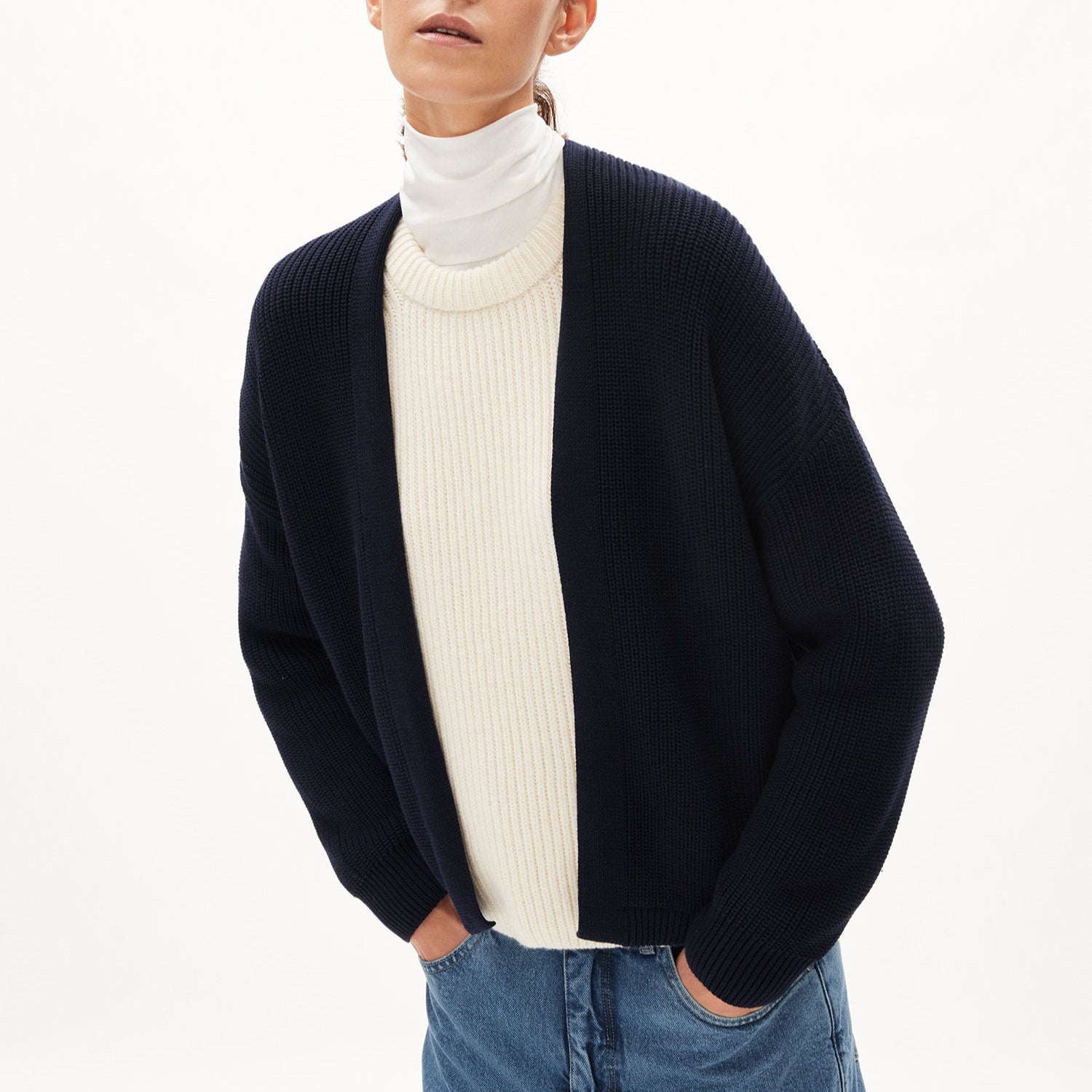 Person trägt eine schwarze Strickjacke über einem weißen Rollkragenpullover und blauen Jeans vor weißem Hintergrund.