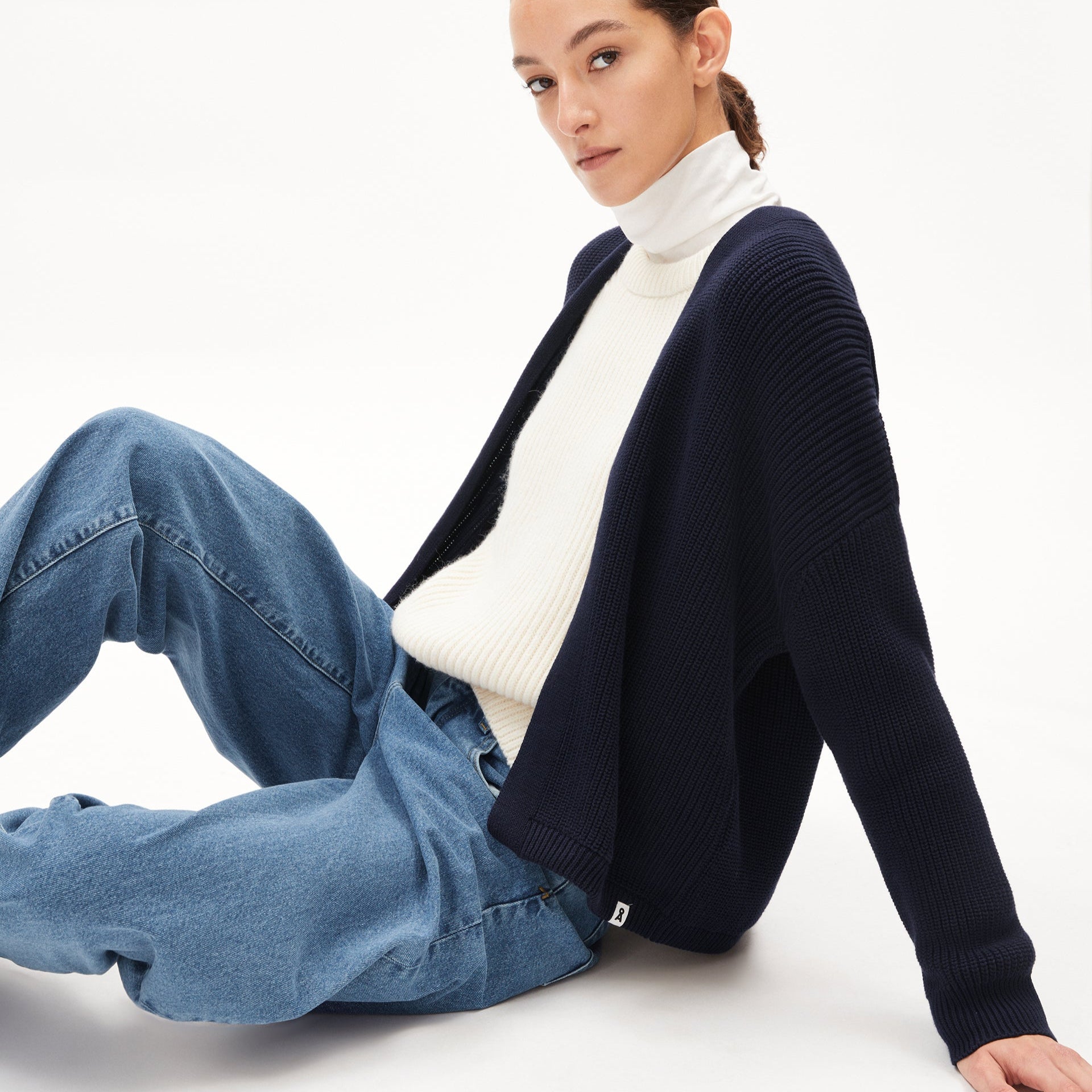 Person sitzt in lässiger Pose, trägt blauen Cardigan, weißen Pullover und Jeans, vor neutralem Hintergrund.