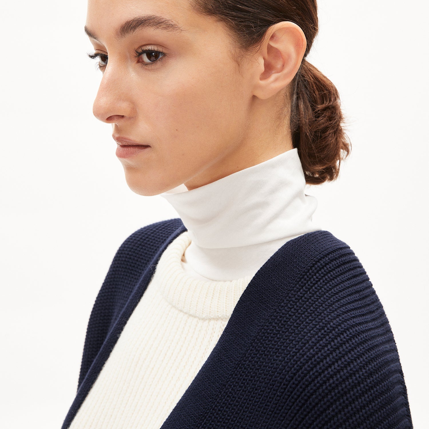 Frau im Profil mit einem weißen Rollkragenpullover und einer blauen Strickjacke vor weißem Hintergrund.