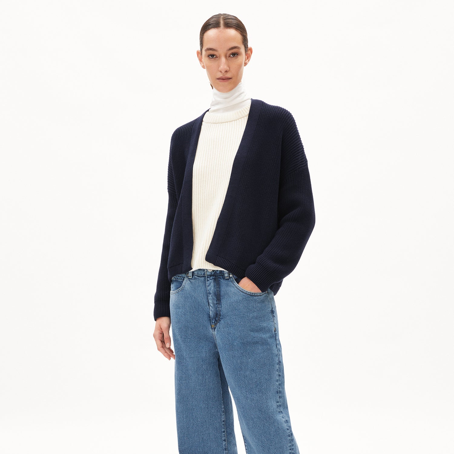 Frau in blauem Cardigan, weißem Rollkragenpullover, und blauer Jeans vor weißem Hintergrund.