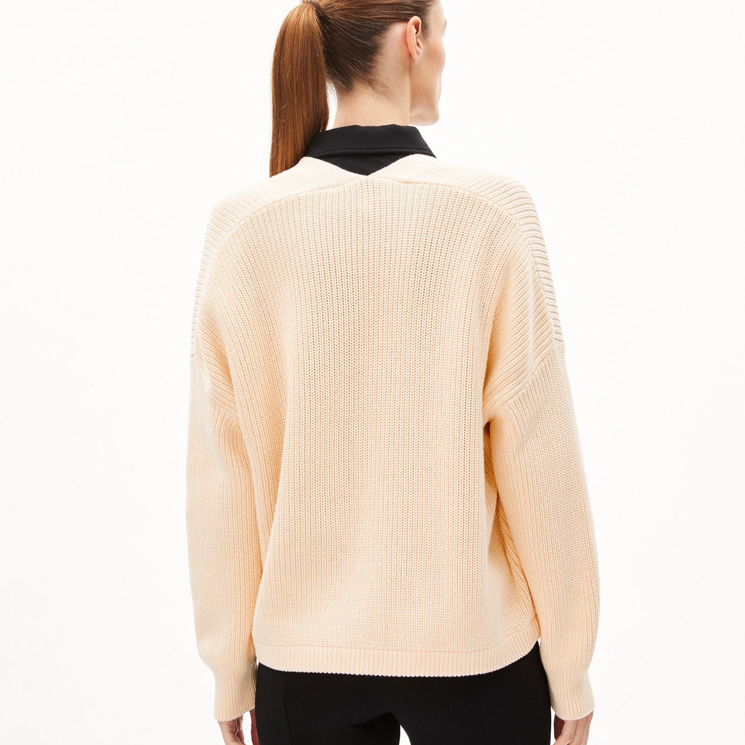 Frau in cremefarbenem Pullover und schwarzer Hose von hinten mit einem seitlichen Pferdeschwanz.