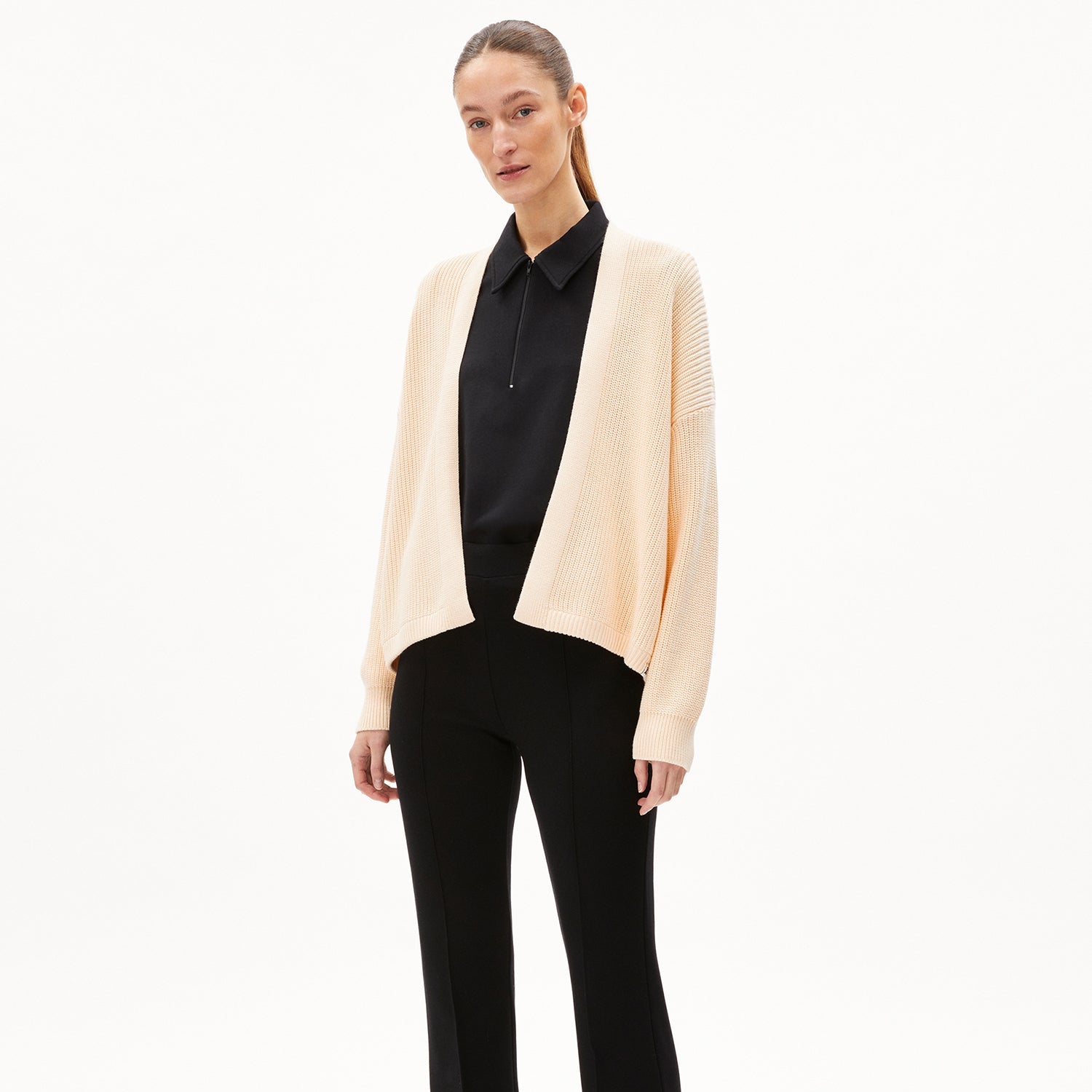 Frau in beige Strickjacke, schwarzem Hemd und schwarzer Hose steht vor einem weißen Hintergrund.