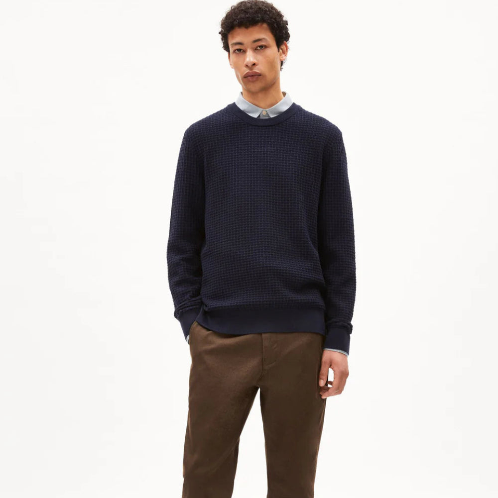 Eine Person steht vor einem schlichten weißen Hintergrund und trägt einen dunkelblauen strukturierten Pullover über einem Hemd mit Kragen, eine braune Hose und graue Turnschuhe. Sie hat kurzes lockiges Haar und einen neutralen Gesichtsausdruck.