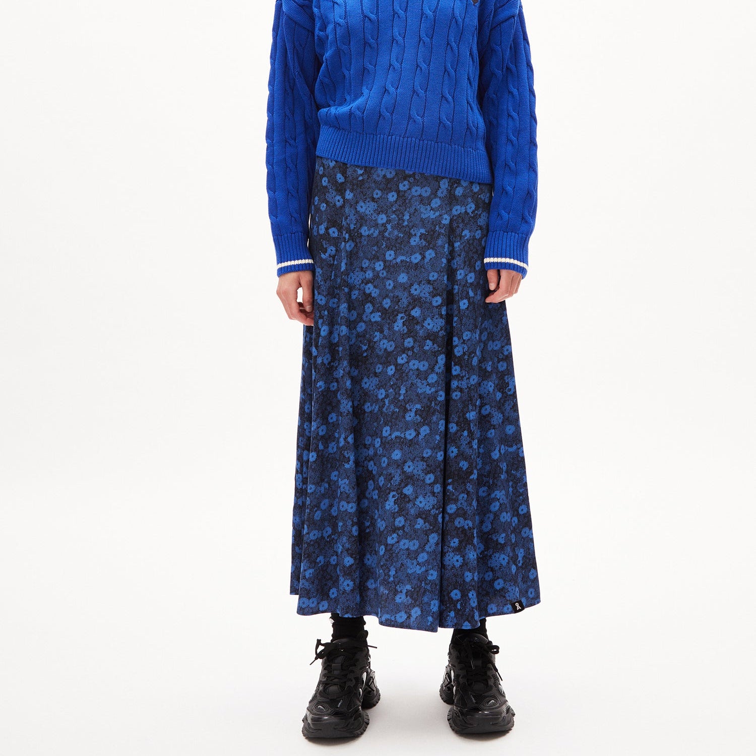 Eine Person trägt einen blauen Pullover und einen langen, blau gemusterten Rock mit schwarzen Schuhen.