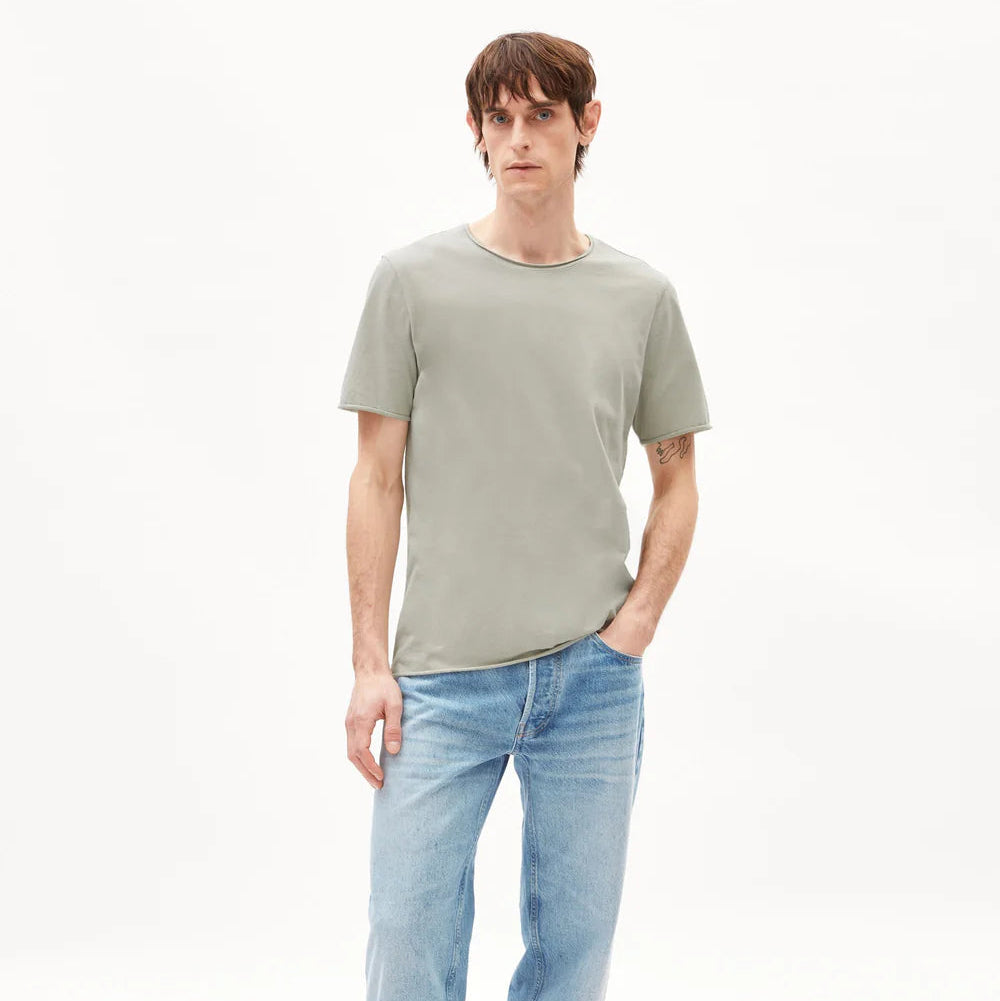 Eine Person steht vor einem schlichten weißen Hintergrund, trägt ein hellgraues, kurzärmeliges T-Shirt, hellblaue Jeans und beigefarbene Halbschuhe und hat eine Hand in der Tasche.