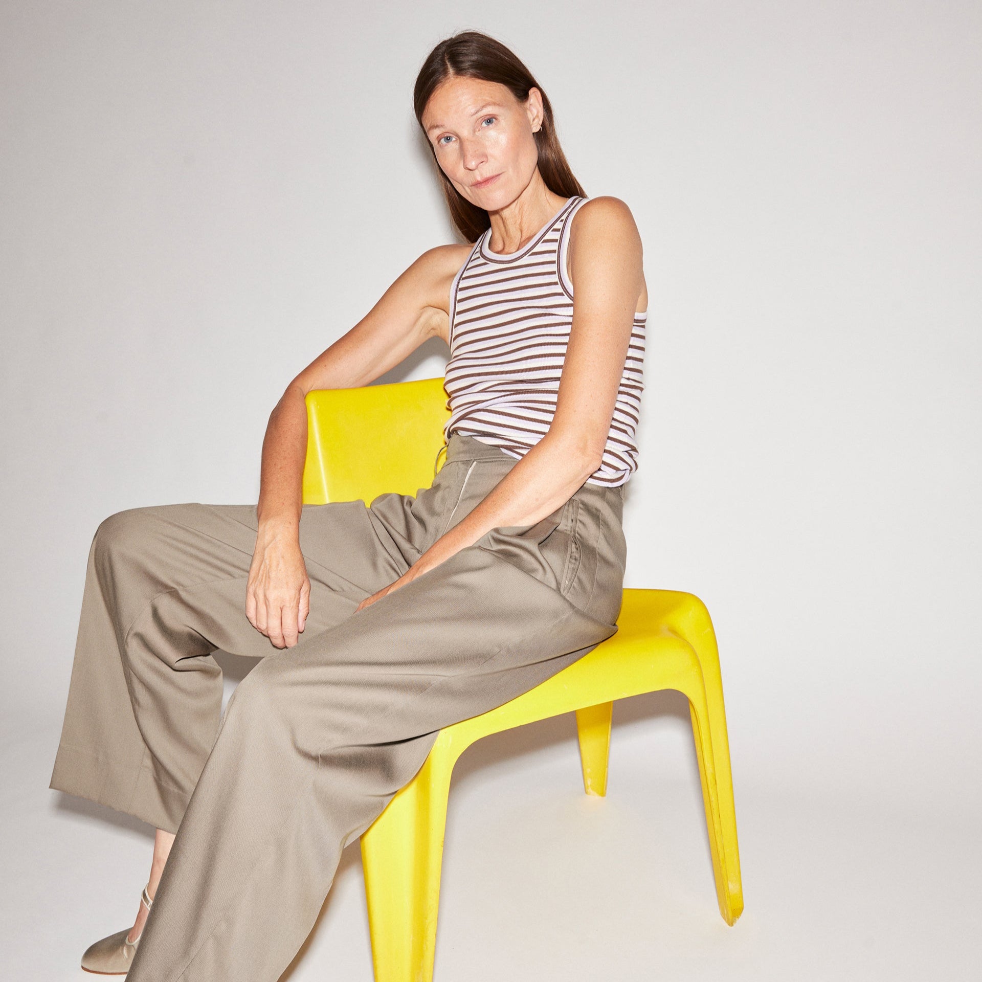 Frau in gestreiftem Oberteil und beiger Hose sitzt auf gelbem Stuhl vor grauem Hintergrund.