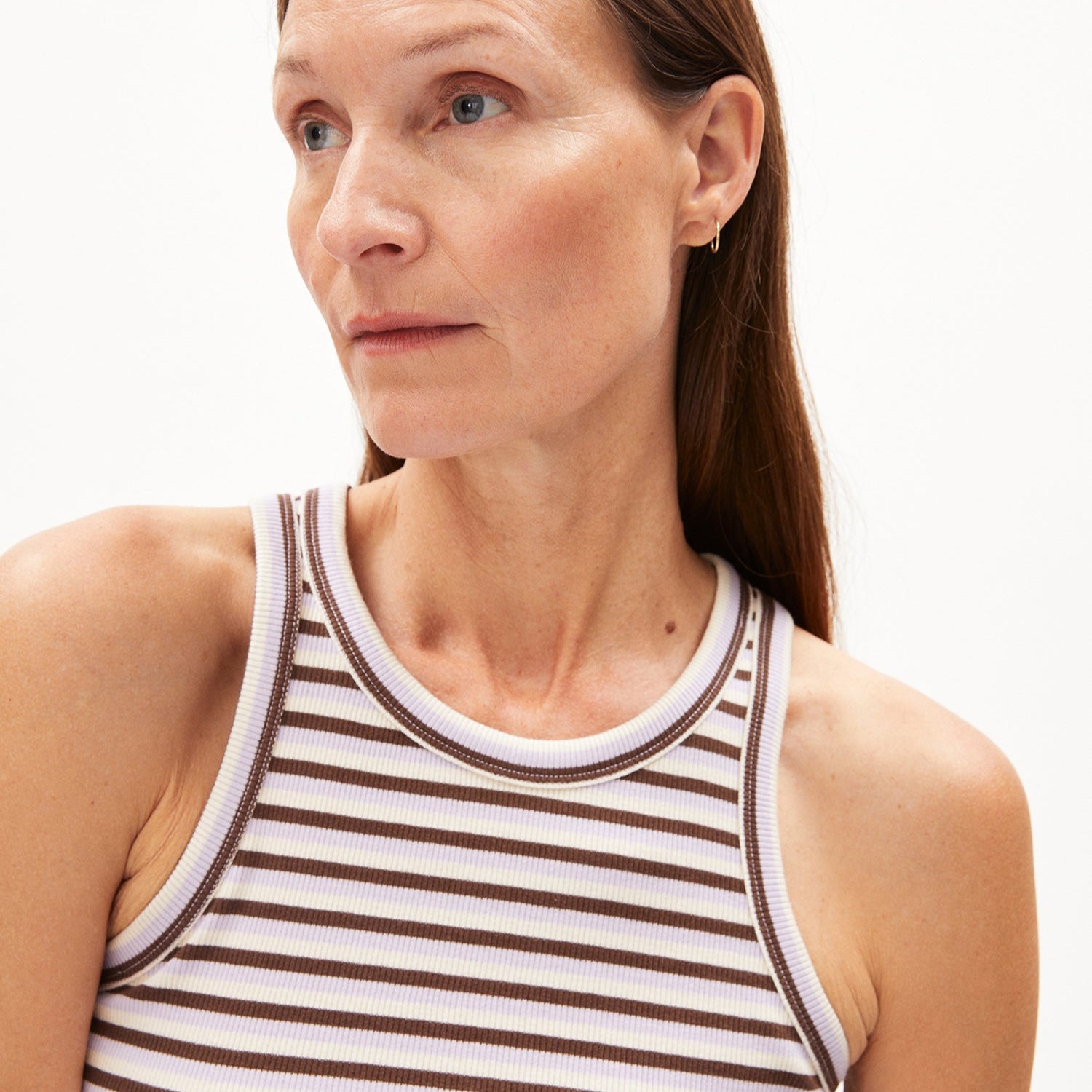 Frau mit langen Haaren trägt ein gestreiftes Tanktop und goldene Ohrringe, schaut nach links gegen einen weißen Hintergrund.