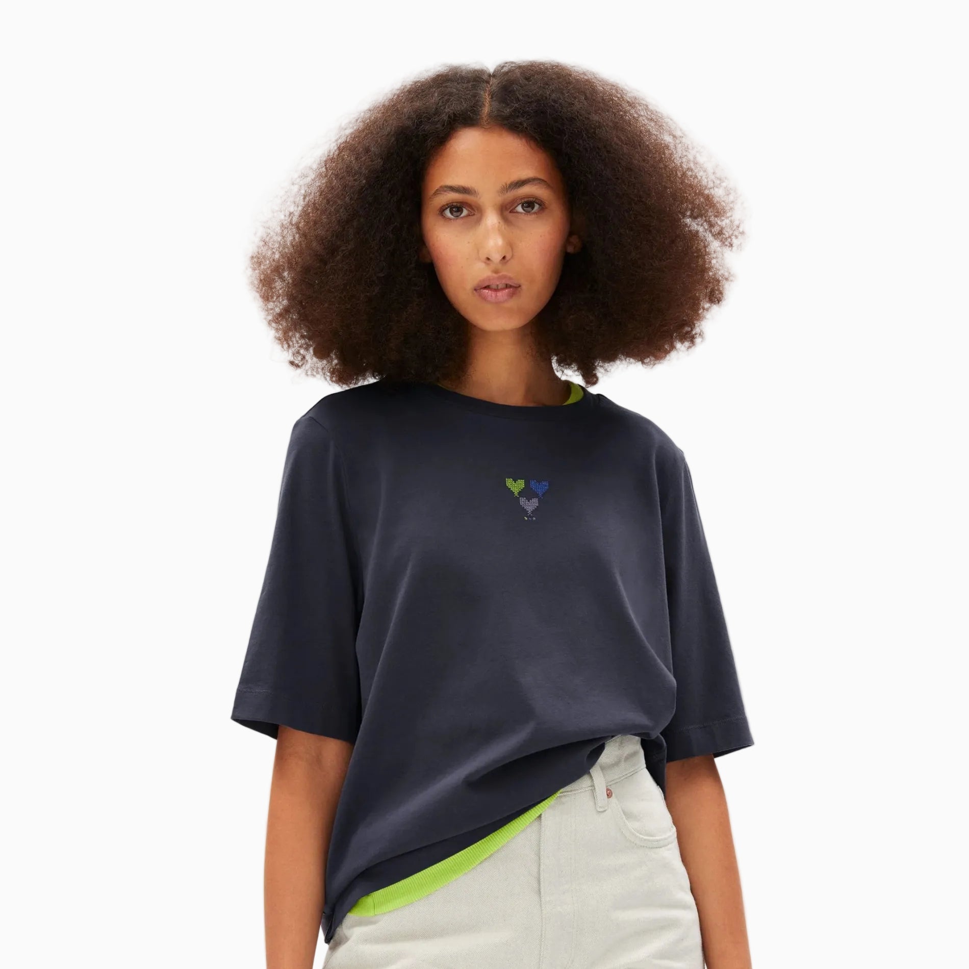 Eine Frau mit voluminösem, lockigem Haar trägt ein locker sitzendes, dunkles, marineblaues T-Shirt mit drei kleinen Herzformen auf der Brust, eine helle Hose und ein angedeutetes neongrünes Hemd, das darunter hervorschaut. Sie steht vor einem schlichten weißen Hintergrund.