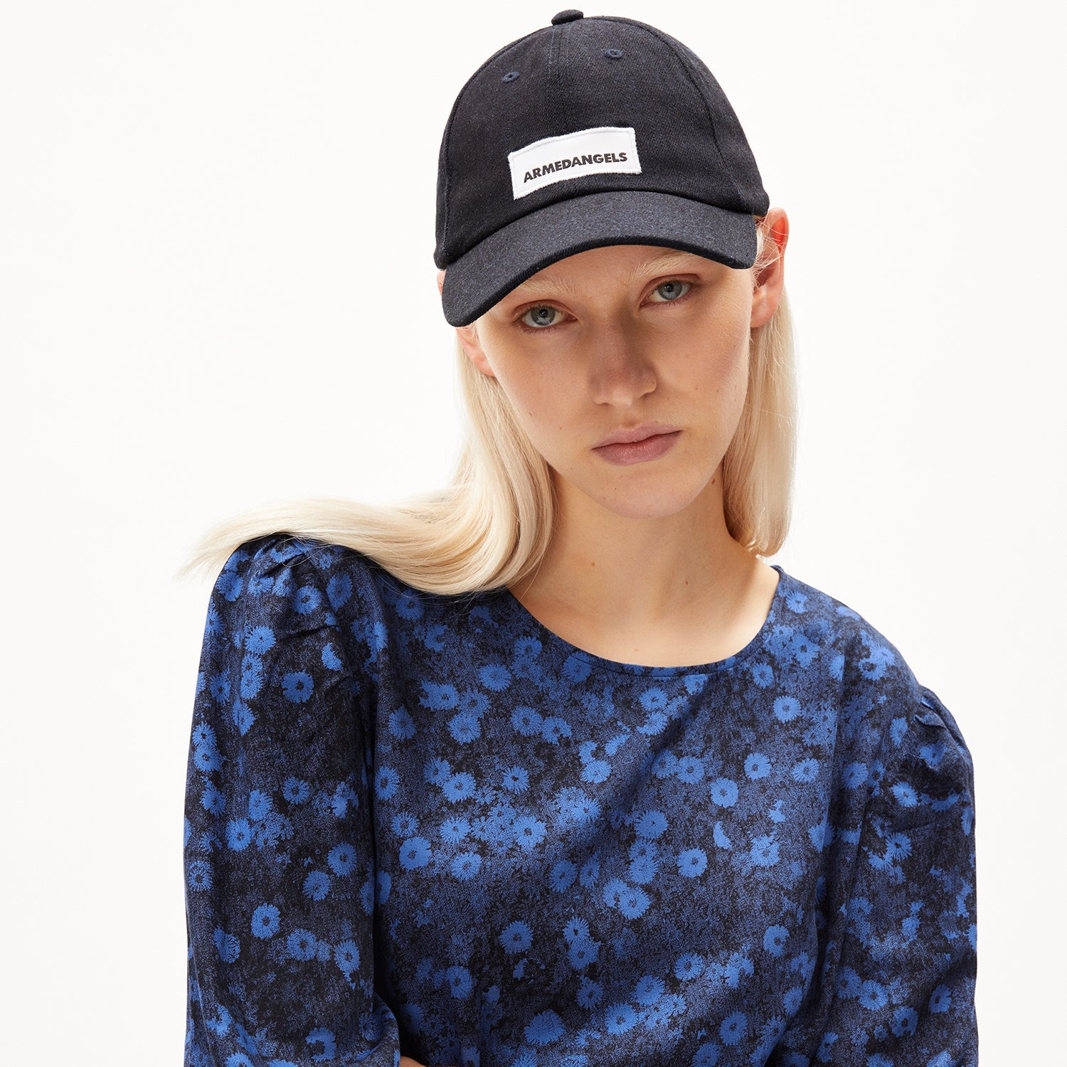 Frau mit blonden Haaren, trägt eine schwarze Kappe mit "ARMDANGELS" und ein blau-blumiges Oberteil, blickt ernst.