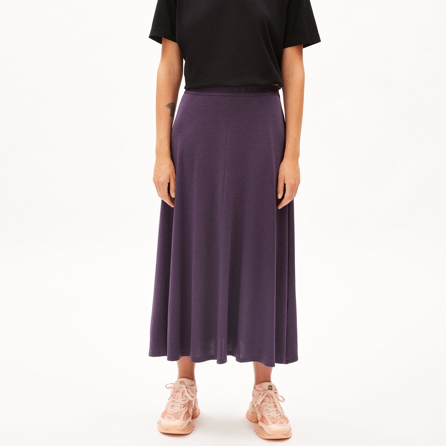 Person trägt einen langen violetten Rock, ein schwarzes T-Shirt und rosa Turnschuhe vor weißem Hintergrund.