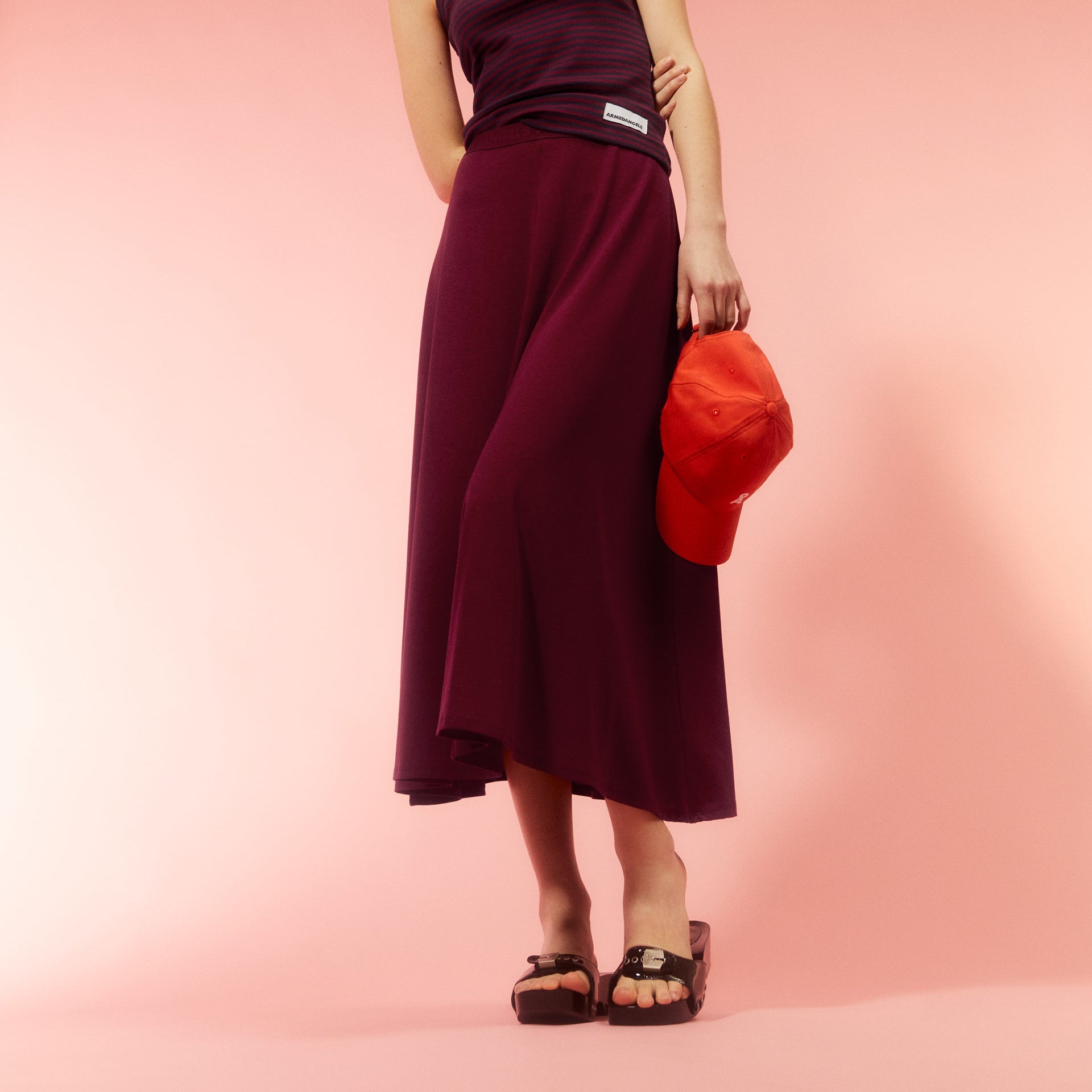 Person in a burgundy skirt and striped top holds an orange cap against a pink background, wearing black sandals.