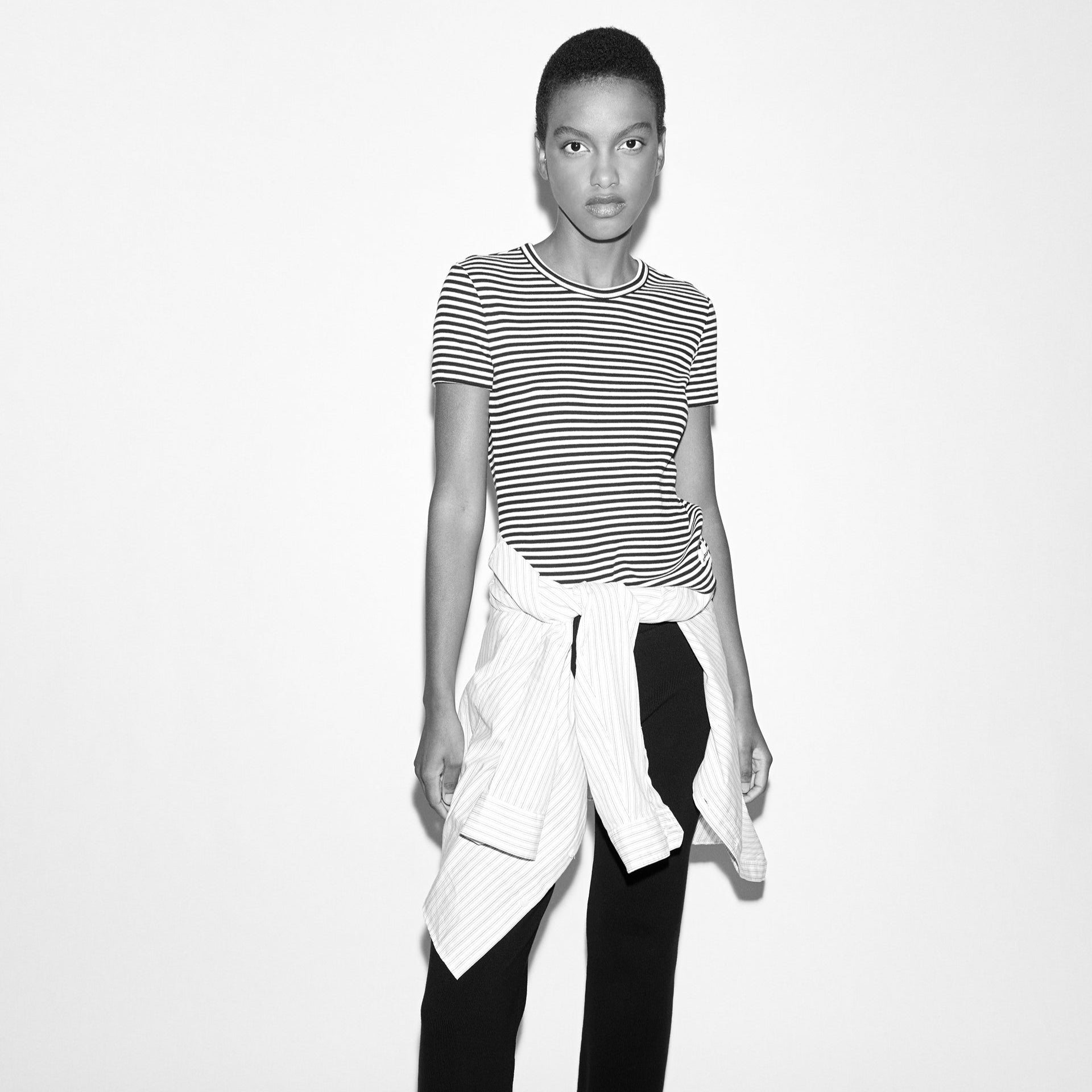 Person in striped shirt and black pants with a shirt tied around the waist, posing against a plain background.