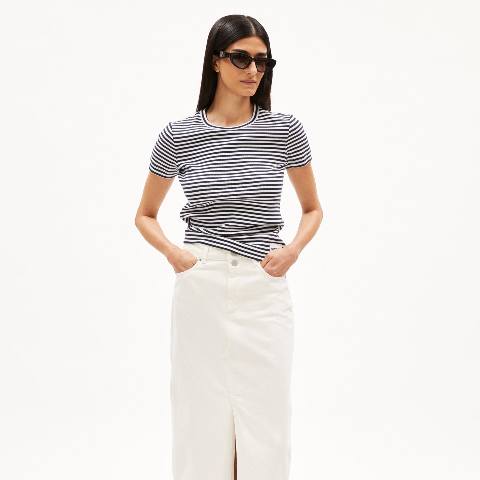 Person with long dark hair wearing sunglasses, a striped black and white shirt, and a white skirt, hands in pockets.