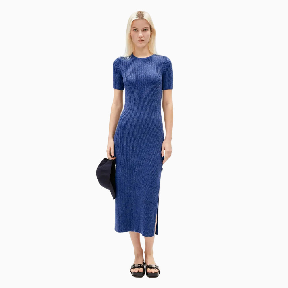 Eine Frau mit langen blonden Haaren trägt ein tailliertes, kurzärmeliges, blaues, geripptes Midikleid mit Seitenschlitzen. Sie hält eine dunkle Kappe in einer Hand und trägt schwarze Sandalen. Sie steht vor einem schlichten weißen Hintergrund.
