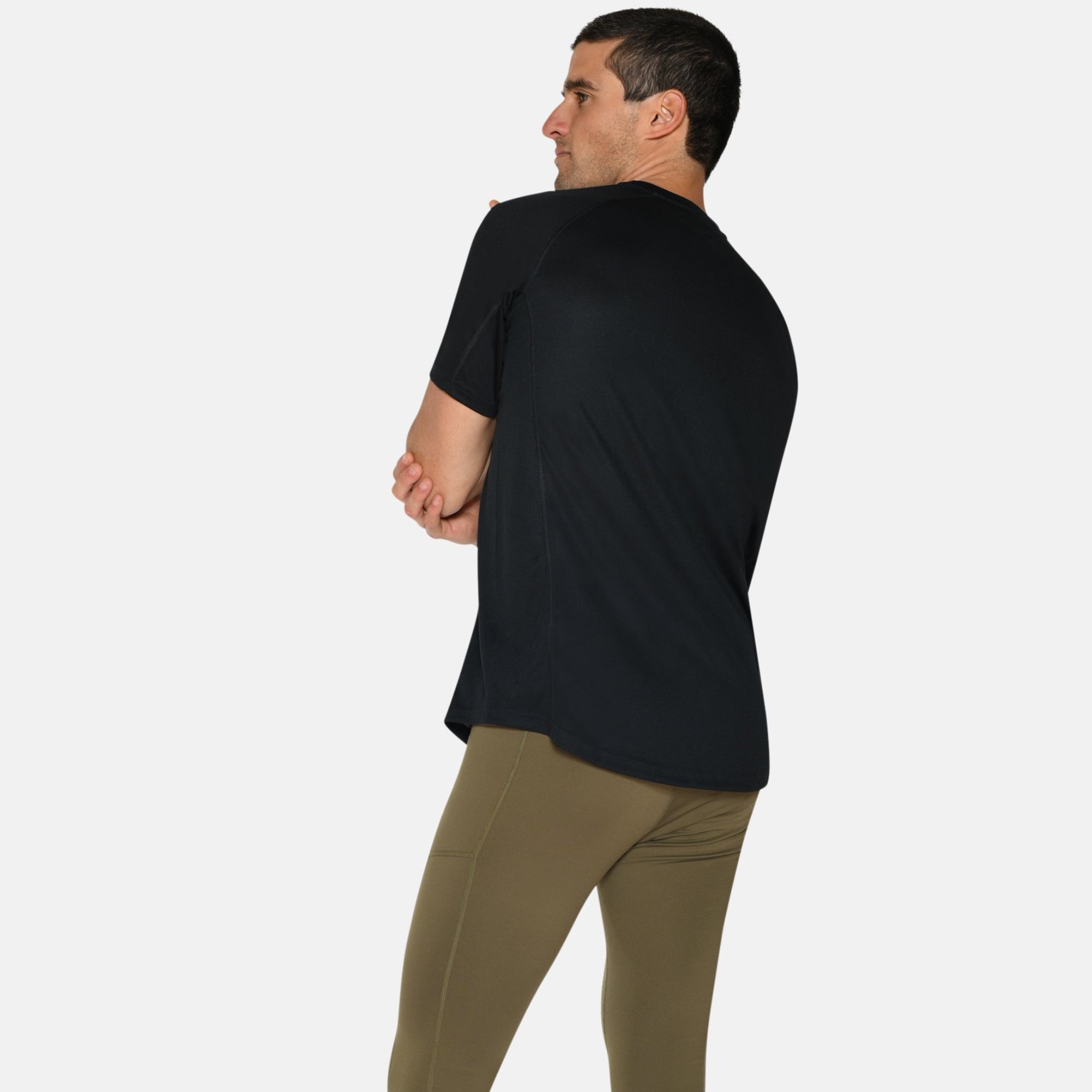 Ein Mann steht mit verschränkten Armen, den Blick abgewandt. Er trägt ein schwarzes T-Shirt, olivgrüne Leggings und blaue Sportschuhe mit orangefarbenen und weißen Akzenten. Der Hintergrund ist einfarbig weiß.