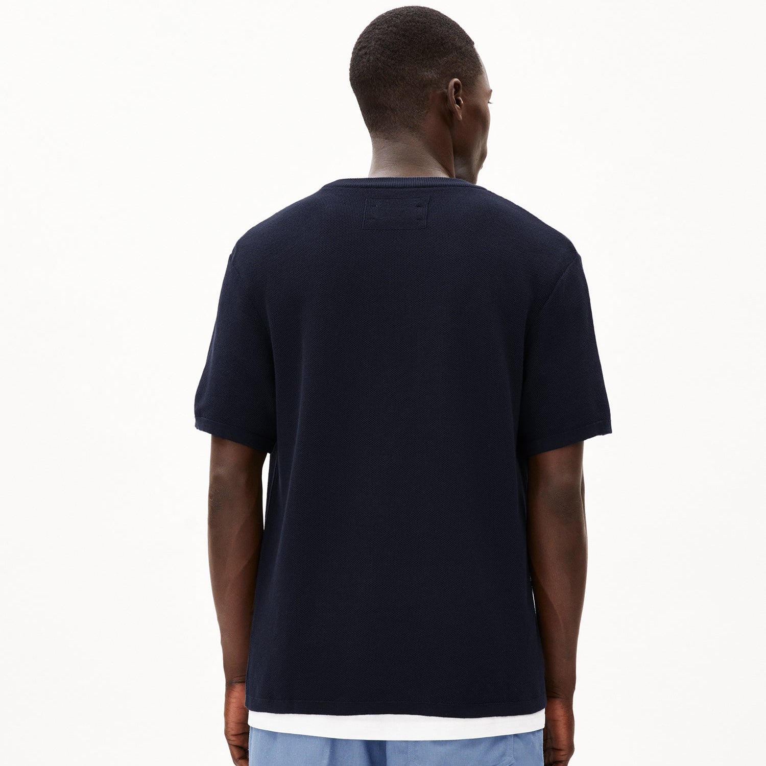 Person von hinten in dunklem T-Shirt und blauer Hose vor weißem Hintergrund stehend.