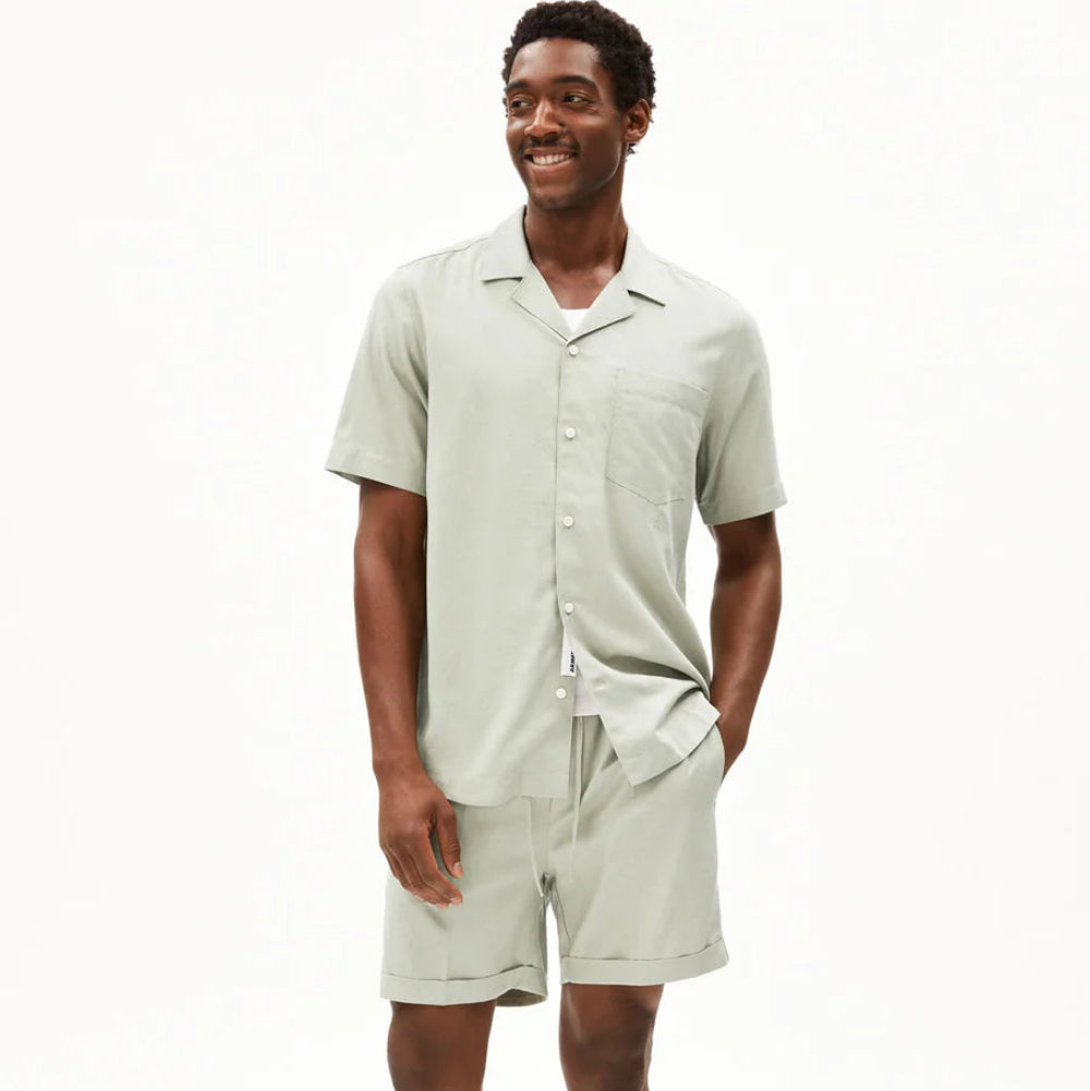 Ein Mann steht lächelnd in einem hellgrünen, kurzärmeligen Button-up-Hemd, passenden Shorts und braunen Halbschuhen. Der Hintergrund ist einfarbig weiß.